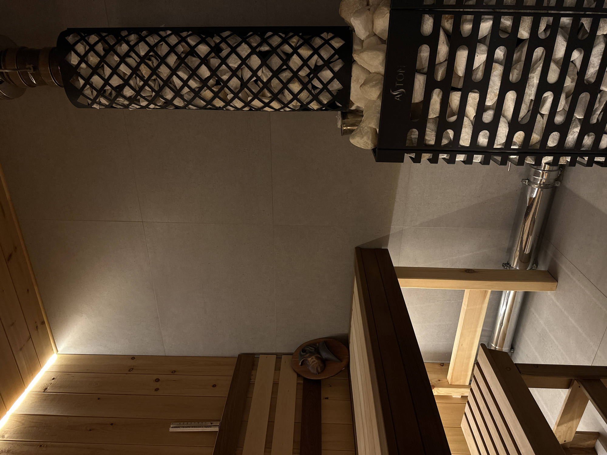 The image depicts the interior of a sauna, possibly located in Прикамский, Russia. Two different sauna stoves filled with rocks are visible at the top of the frame, one cylindrical with a black metal cage and another rectangular, also with a black metal frame and the brand "ASTON" visible on it. A metal chimney extends from both stoves. The floor and wall are tiled with large, light gray tiles. In the lower portion of the image, there is a wooden structure with what appears to be sauna benches, constructed of light-colored wood. A round, wooden decoration is also visible, mounted on a section of the wood paneling. An LED strip light is also visible in the sauna.