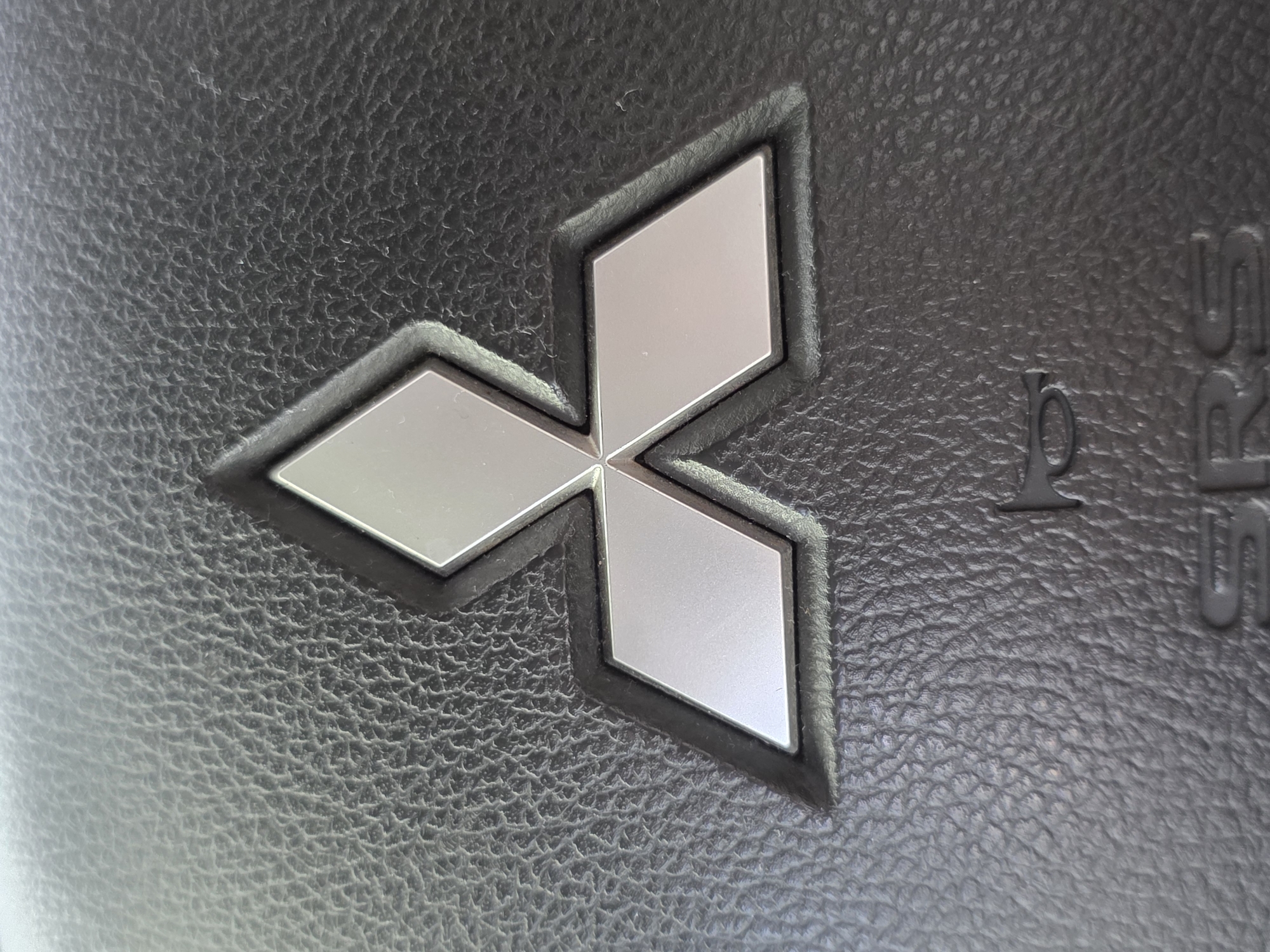 The image shows a close-up of a car's steering wheel, featuring the Mitsubishi logo. The logo is made of three silver diamond shapes arranged in a triangular pattern, set against a black, textured background that appears to be leather. Below the logo, the outline of a horn symbol is visible, followed by the embossed letters "SRS," which indicates an airbag system. The overall scene is a static, close-up shot of an interior car component. There is no information in the image to indicate its location in Chaikovsky, Russia.