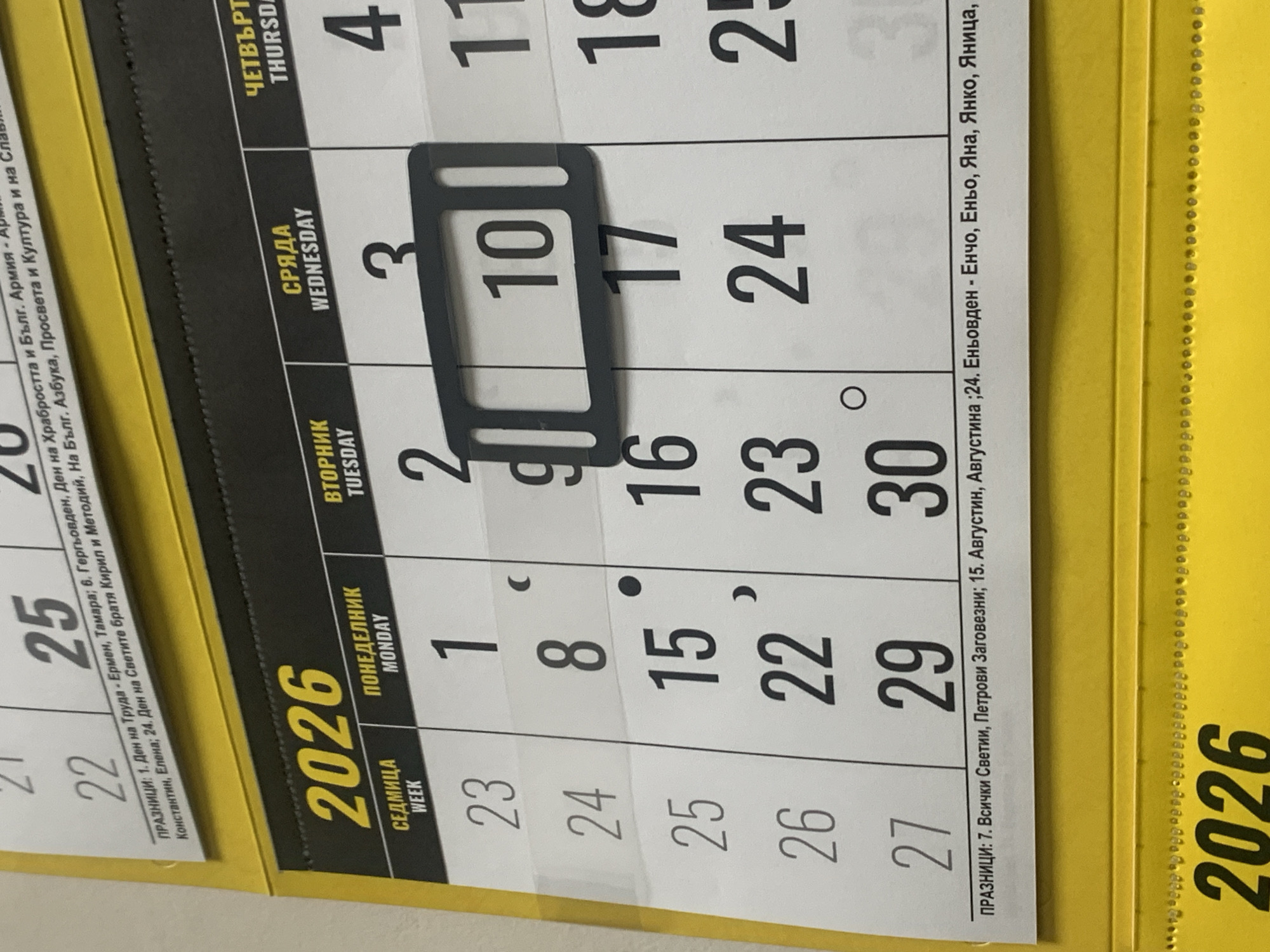 The image displays a portion of a calendar for the year 2026. The calendar is divided into days of the week and dates. Prominently visible are the numbers 2026, and specific dates such as 1, 2, 3, 4, 8, 9, 10, 11, 15, 16, 17, 18, 22, 23, 24, 25, 26, 27, 29, 30. There is also text in Bulgarian indicating weekdays like "ПОНЕДЕЛНИК" (Monday), "ВТОРНИК" (Tuesday), "СРЯДА" (Wednesday), and "ЧЕТВЪРТЪК" (Thursday), along with their English translations. Some dates are marked with small dots and commas, likely indicating holidays or special events. The calendar appears to be hanging on a wall, with a yellow border at the top and bottom. The overall context suggests a typical office or home setting where a calendar would be displayed. There is no visual information in the image that allows for the determination of the specific location within the city of Burgas, Bulgaria, or any other specific geographical location.