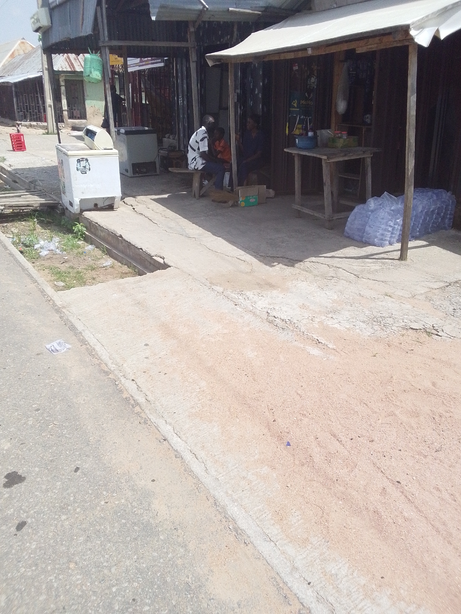 The image depicts a street scene in Jalingo, Nigeria, likely outside a small market stall. In the foreground, a paved road transitions into a concrete walkway bordering a drainage ditch filled with grass and debris. To the left, a white freezer sits on the walkway, with a red plastic crate visible behind it. Further back, under a corrugated metal awning, two people are seated on a wooden bench. One person wears a white and black striped shirt and blue trousers, while the other wears an orange shirt and blue trousers. A wooden table is positioned in front of them, displaying a blue bowl and other items. To the right of the table, stacks of bottled water are piled on the ground. Behind the stall, structures with corrugated metal roofs and barred windows are visible. The environment appears to be a casual outdoor commercial area with signs of everyday life and commercial activity.