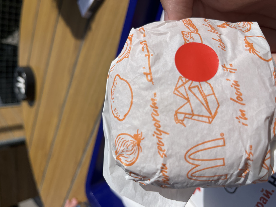The image shows a hand holding a McDonald's hamburger wrapped in paper. The paper is white with orange and red illustrations. The hand is holding the bottom of the hamburger. The image was taken in the city of Hadjidimitrovo, Bulgaria. The location is a restaurant with a wooden table and a blue tray. There is a cigarette butt container in the background, blurred. The paper has different languages, including Turkish, Arabic, and English. The English text on the paper reads "I'm lovin' it". The hamburger has a red sticker, a McDonald's logo, and other illustrations, including a lemon, onion, and a crane. A blue container is in the background. The photo was taken during the day. The weather is sunny.