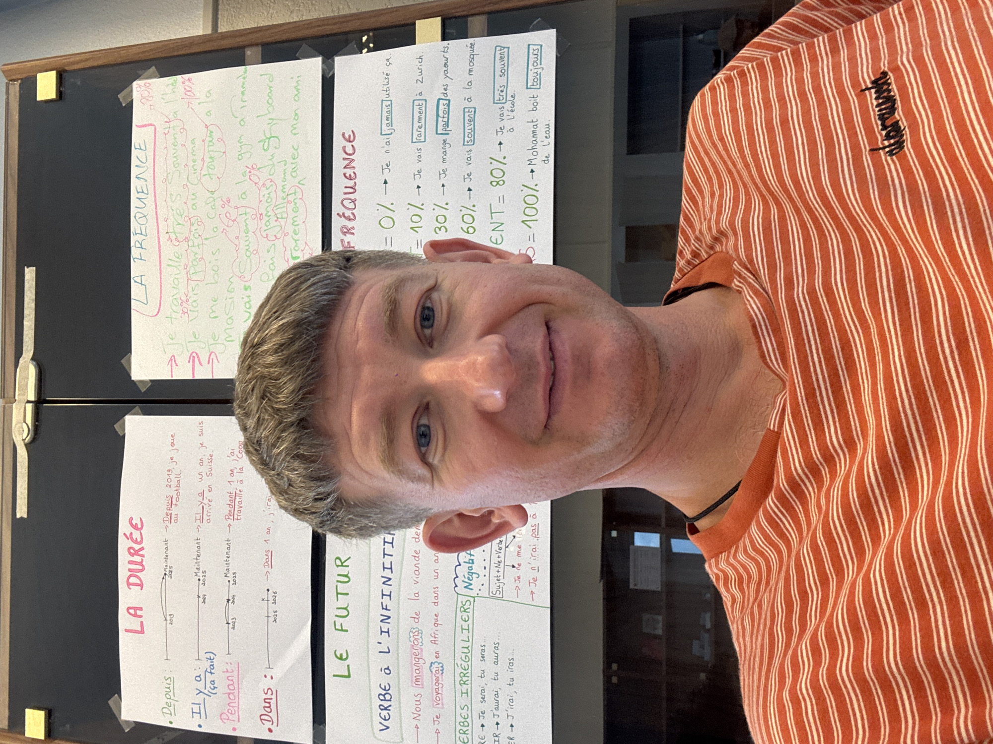 The image shows a medium close-up shot of a man's face and upper torso, taken outdoors. The man is looking directly at the camera with a slight smile. He is wearing an orange and white striped t-shirt with the word "alprausch" embroidered in black on the left side of his chest.
Behind the man, a bulletin board is visible, covered with several handwritten notes in French. These notes appear to be educational materials, possibly for a language class. One section is titled "LA FRÉQUENCE" (Frequency) and includes percentages and phrases related to how often actions occur. Another section is labeled "LA DURÉE" (Duration) and features a timeline with dates and events. A third section is titled "LE FUTUR" (The Future) and discusses verbs in the infinitive and irregular verbs.
The lighting suggests it is daytime, but the overall scene appears to be indoors or in a shaded outdoor area, given the quality of light and the presence of the bulletin board. The background is blurred, but hints of a room with shelving or furniture can be seen. The man's expression is friendly and approachable. The presence of the bulletin board and the educational content strongly suggests a school or learning environment. The word "Bellelay" in the prompt provides a specific location context for this scene.