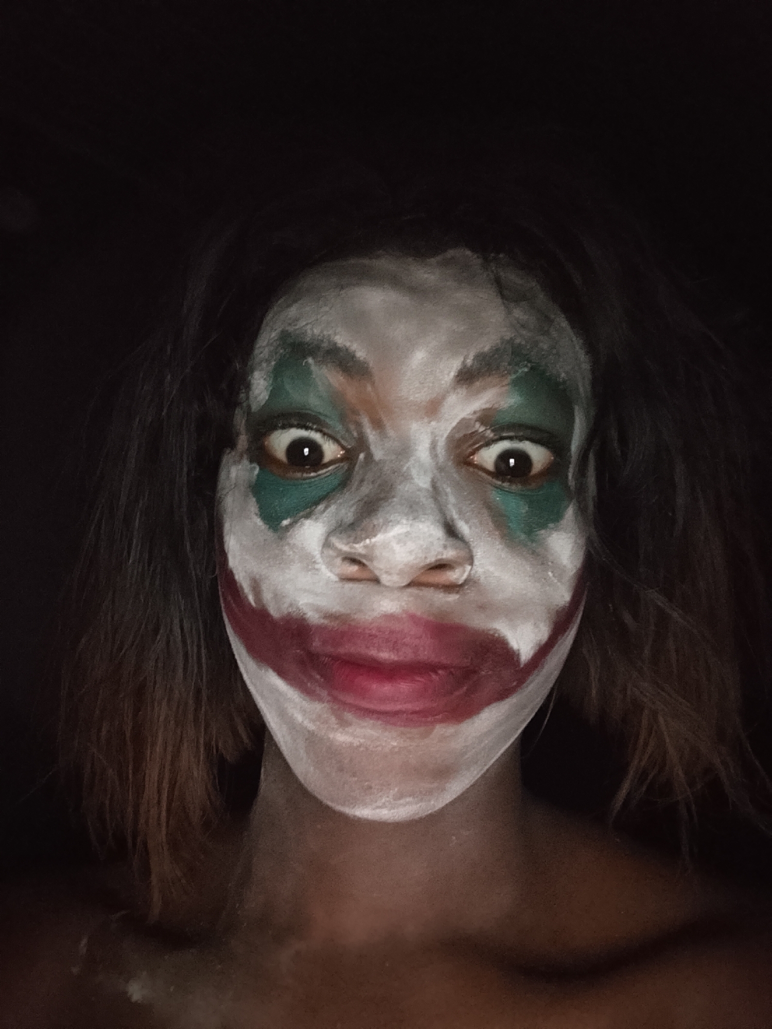 A headshot of a person with smeared white face paint, green paint around the eyes, and red paint around the mouth to resemble the Joker. The background is dark. The location is unknown.