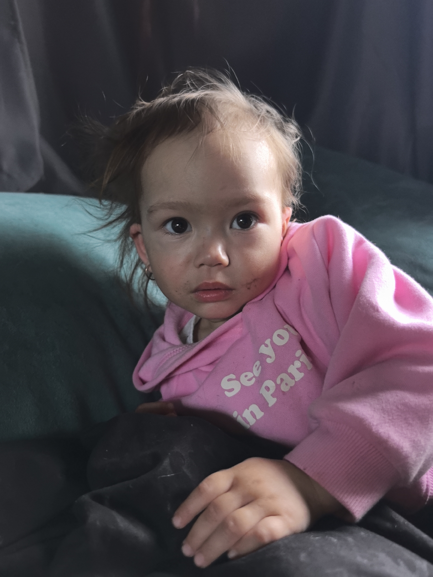 The photograph captures a young toddler in an indoor setting, likely a home in Noviy, Russia. The child, with light brown, slightly disheveled hair and large, dark eyes, looks directly at the camera with a neutral, possibly curious, expression. Dark smudges are visible on the child's forehead, cheeks, and around the mouth, suggesting a recent messy activity. The child wears a bright pink hooded sweatshirt, which has white text on the front partially visible, reading "See you in Pari" likely "See you in Paris". A small gold hoop earring is present in the child's left ear. The lower half of the child's attire consists of dark-colored pants or bottoms. The child is seated on a teal or dark green upholstered surface, possibly a couch. The background is dark and out of focus, appearing as a curtain or wall. The scene depicts a quiet, personal moment. The time of day is not discernible from the indoor lighting.