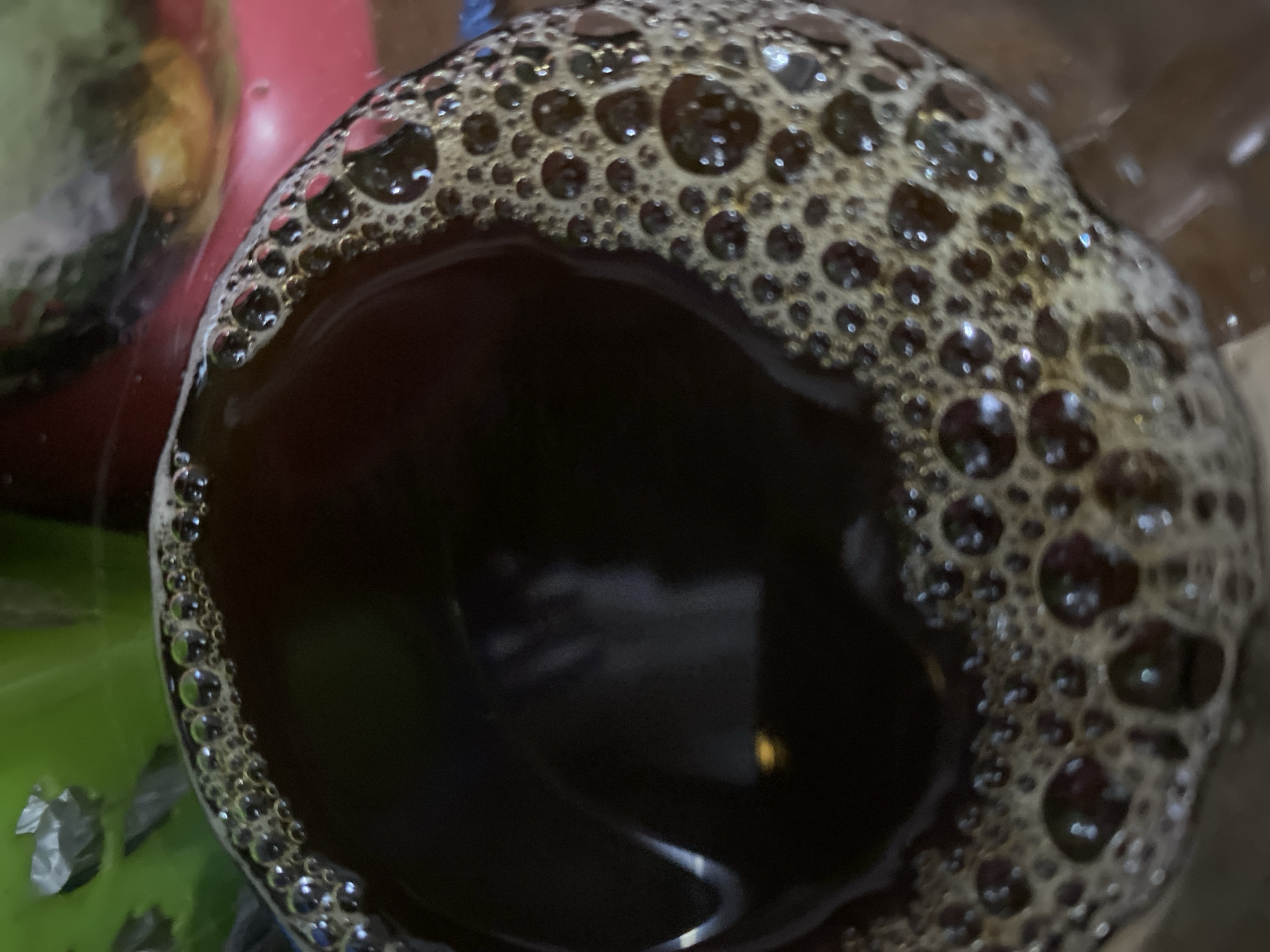 The image shows a close-up, overhead view of a dark, foamy liquid, likely coffee, in a clear container. The foam is composed of numerous small bubbles, with some larger ones interspersed. The liquid itself is a deep brown color, with reflections visible on its surface. The background is out of focus, with hints of green and red objects, suggesting an indoor setting, possibly a kitchen or cafe. There is no discernible location in the city of Kasihan, Indonesia, visible in this image. The image is a still photograph and does not depict any people, actions, or interactions.