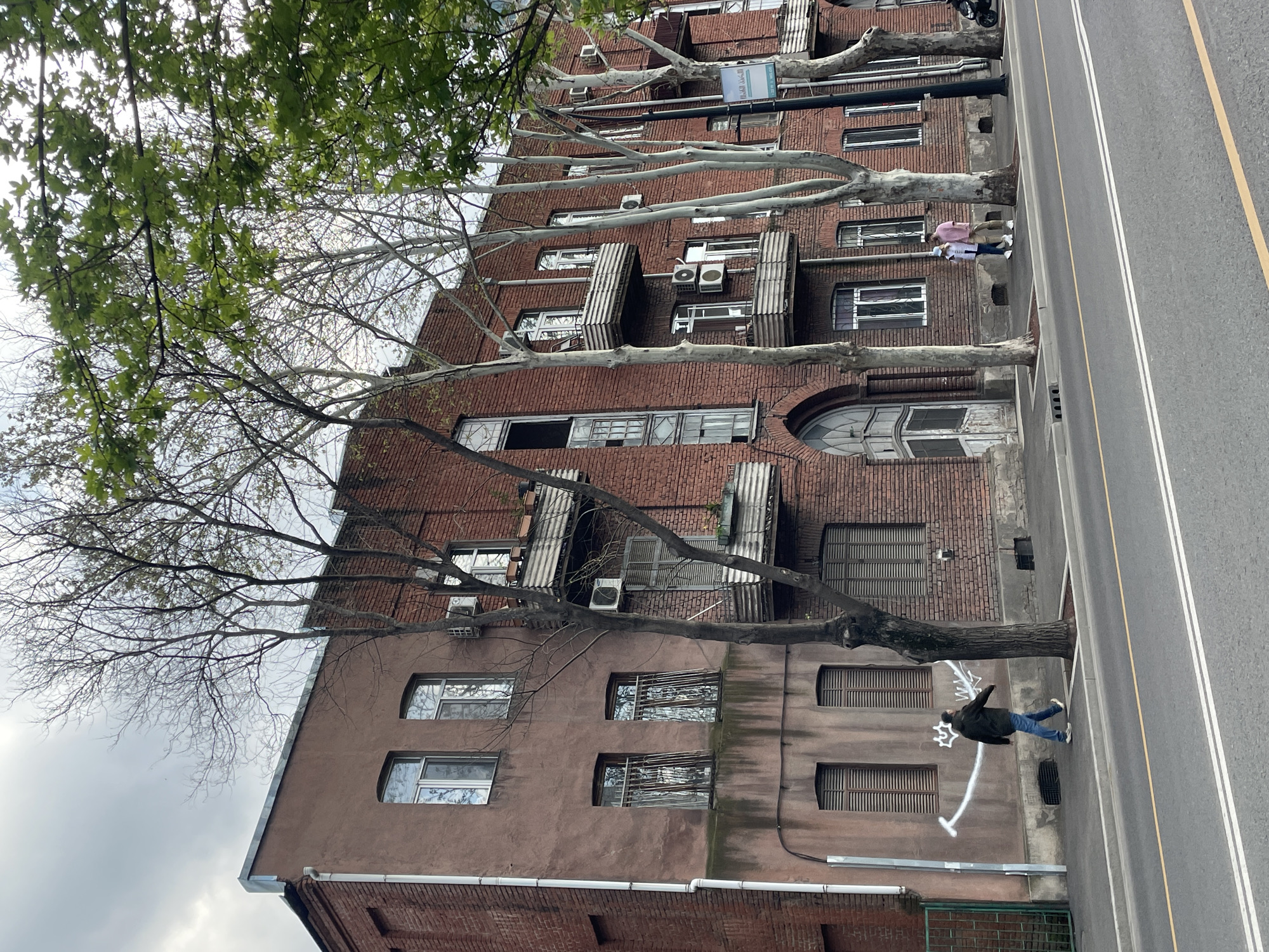 A photograph shows a multi-story building in Tbilisi, Georgia, likely residential or mixed-use, adjacent to a street. The building is constructed primarily of red brick and features a number of windows, some of which have metal grates or bars. Several air conditioning units are mounted on the exterior of the building.
Trees with white bark line the street, their branches extending towards the building. A few people are visible: one person is walking on the street, their clothing is a pink top and khaki pants. Another person walks further down the street, wearing a dark jacket and blue jeans. White graffiti is visible on the building next to the person in the dark jacket and blue jeans.
The street is paved and includes white lines. The sky is overcast, indicating the photograph was taken on a cloudy day.