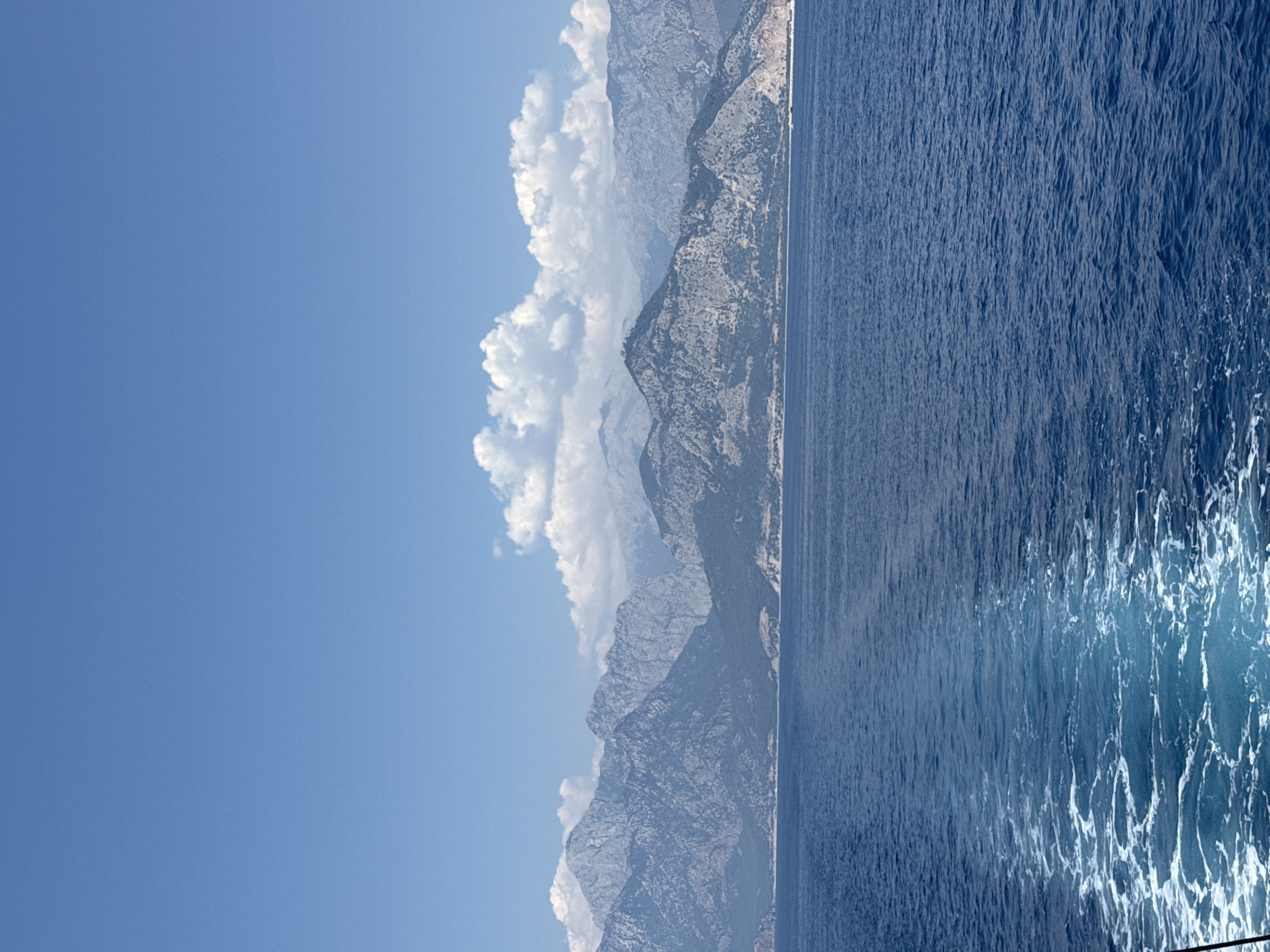 This is a vertical, low-angle, outdoor, daylight photograph showing a scenic view of the Mediterranean Sea and mountains. The image is dominated by a vast expanse of deep blue water on the right, with white foam and ripples suggesting movement, perhaps from a boat. The left side of the image features a majestic mountain range with rugged, rocky slopes, partially obscured by dramatic, puffy white clouds. The sky above is a clear, vibrant blue. The mountains appear dry and sparsely vegetated, with visible textures of rock formations. The light suggests it is daytime, likely late morning or early afternoon, given the brightness and the shadows on the mountains. The overall impression is one of natural beauty and tranquility. The location is identified as Konyaaltı, Turkey, which is known for its coastline and mountainous backdrop. There is no visible text in the image.