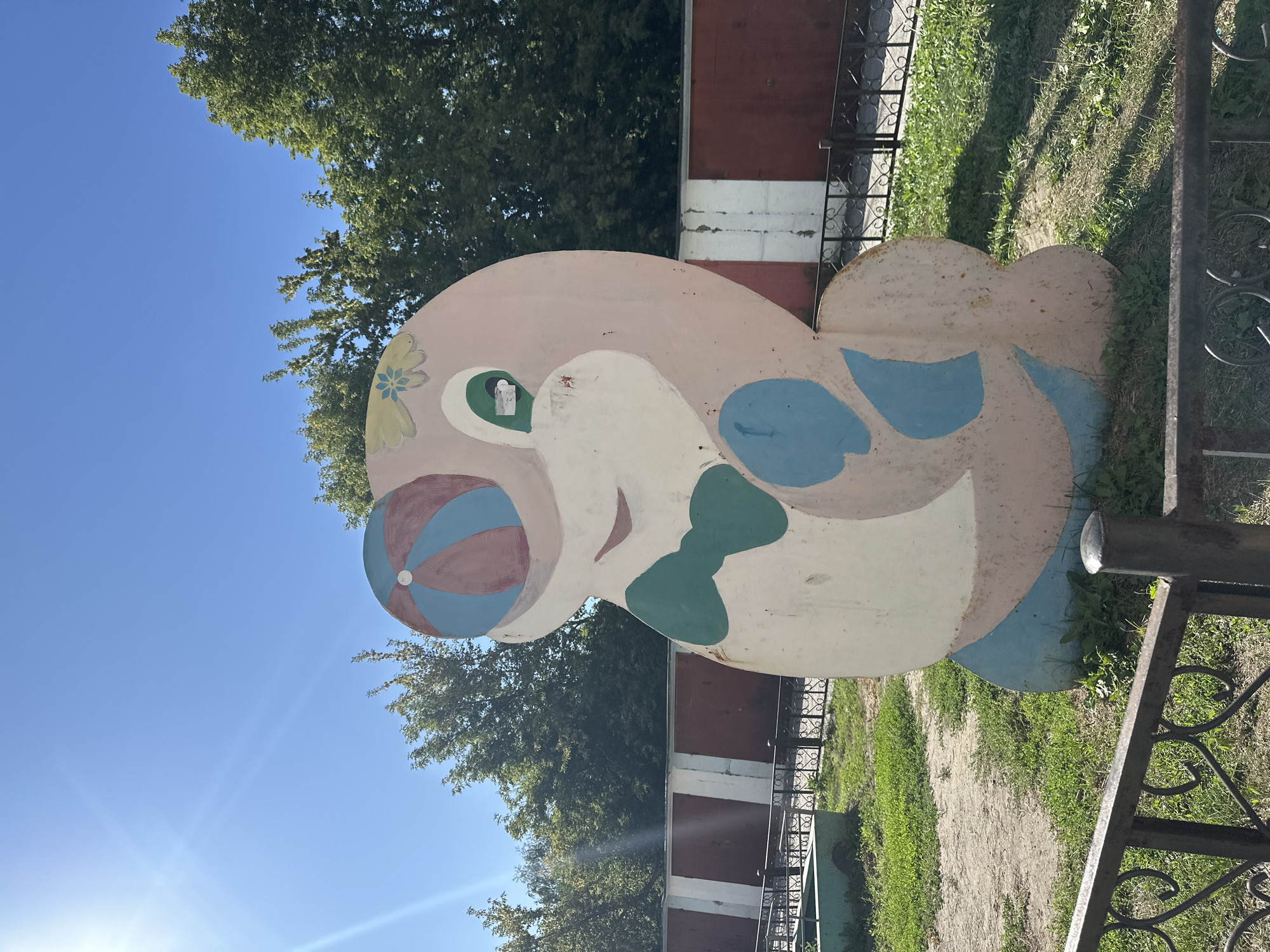 This is an outdoor, eye-level, medium shot of a faded, pink, cartoonish sculpture of a seal, painted with a green bowtie and a colorful ball on its head, standing in a grassy area. The sculpture is located in front of a reddish-brown and white striped wall with a black metal fence in the foreground. Trees with green foliage are visible in the background. The sky is clear and blue with a sun flare on the left side of the image. The sculpture is a likely remnant of a children's playground or recreational area. The location is identified as being in the city of Biysk, Russia.
