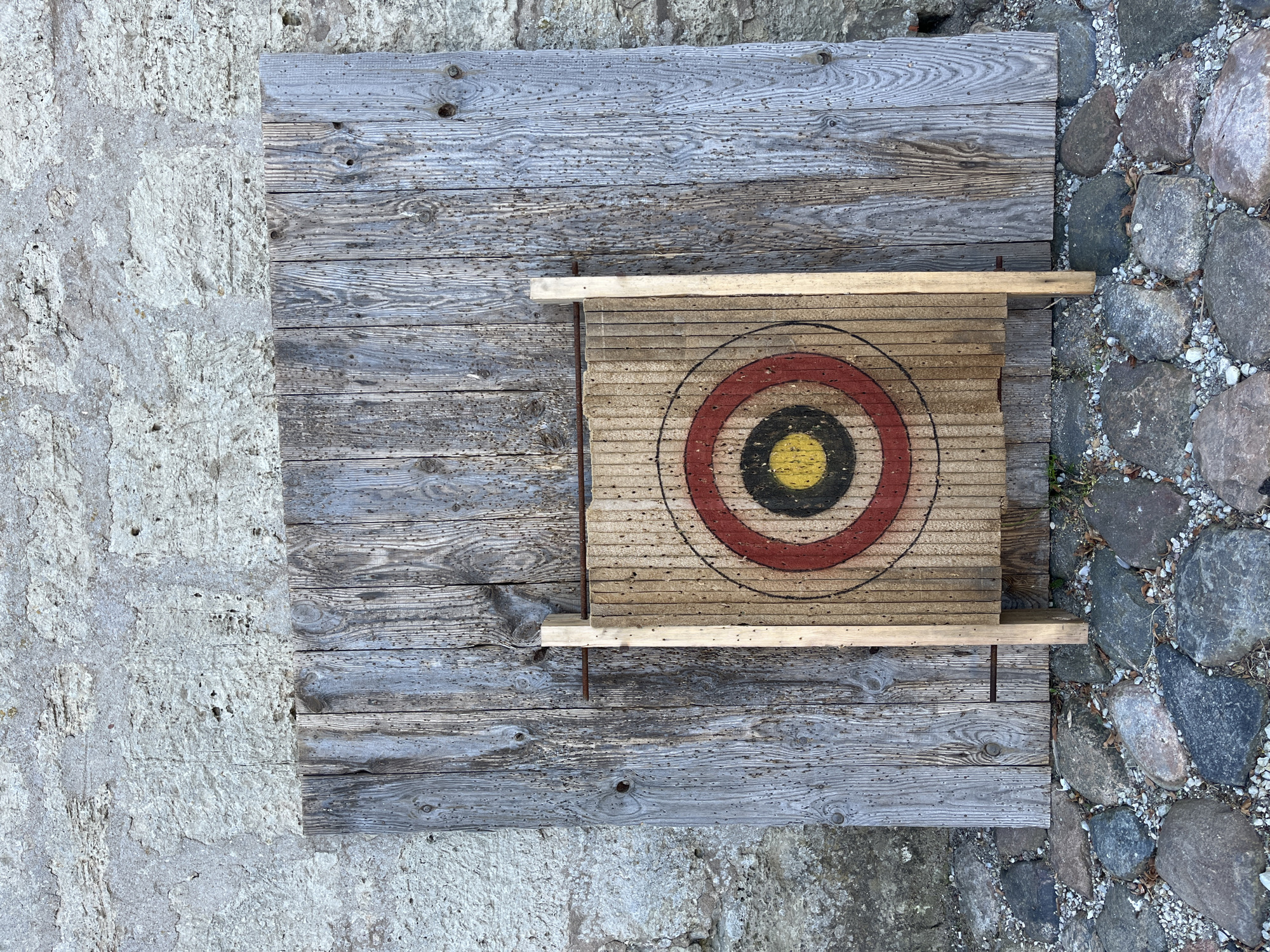 The image depicts a target with concentric rings of red, black, and yellow, painted on a wooden board. This target is propped against a larger, weathered wooden panel, which is itself leaning against a rough, textured stone wall. The ground in front of the structure is covered with a mix of large, grey stones and smaller gravel. The scene appears to be outdoors in a rustic setting, possibly a courtyard or a rural area. Based on the provided context, this scene is located in Kuressaare, Estonia.