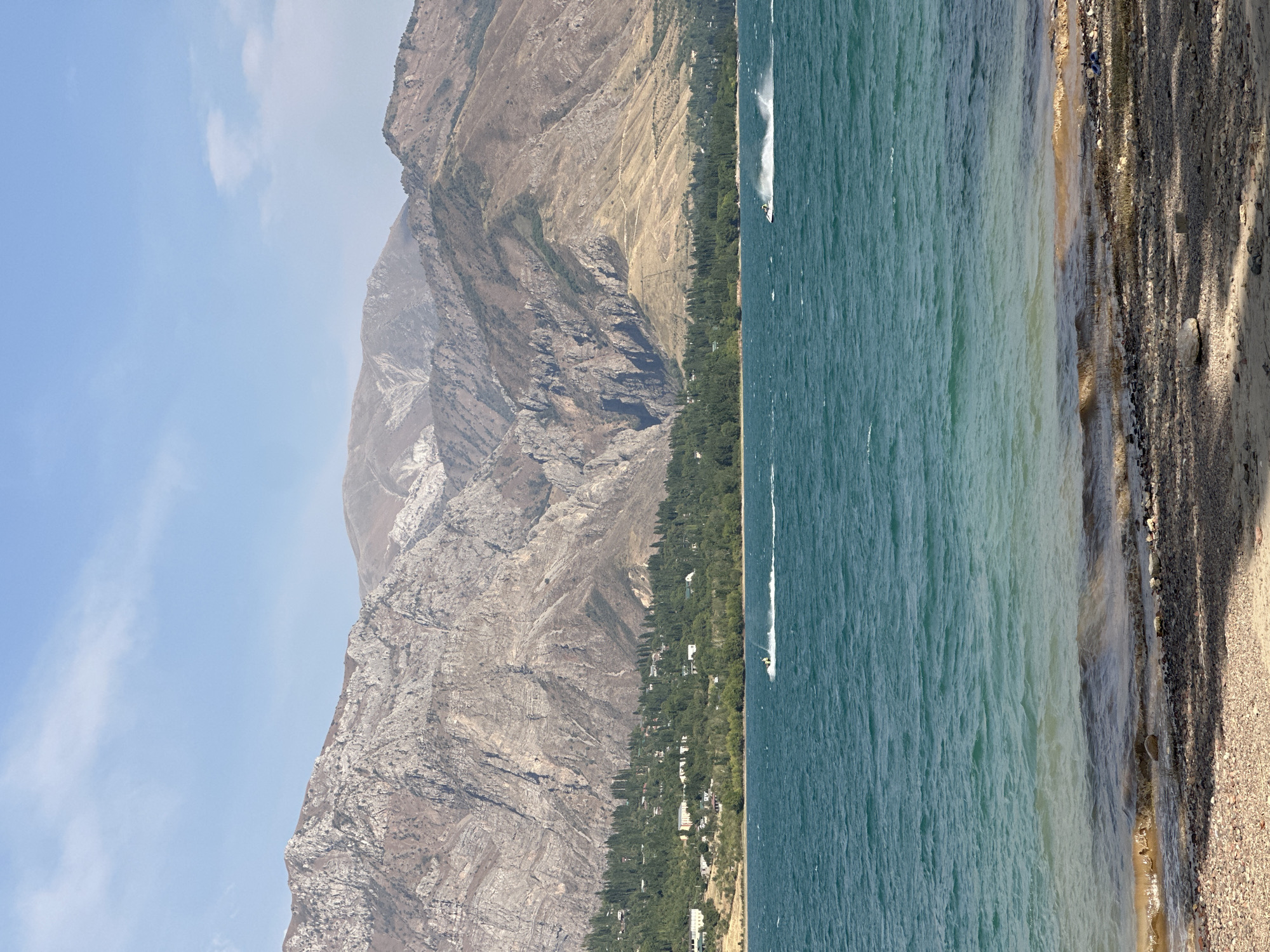 The image depicts a wide, tranquil body of water with ripples and waves under a hazy blue sky. In the distance, a lush green treeline runs along the shore, dotted with houses and buildings. Behind the trees, imposing, rugged mountains rise up, their rocky surfaces marked by shadows and sunlight. The foreground shows a shoreline composed of sand and pebbles. Two individuals on jet skis are visible on the water, creating white wakes as they move. The scene appears to be a recreational area, possibly a lake or reservoir near a mountainous region, fitting the description of a location within Yusuphana, Uzbekistan, which is known for its natural beauty and proximity to mountains.