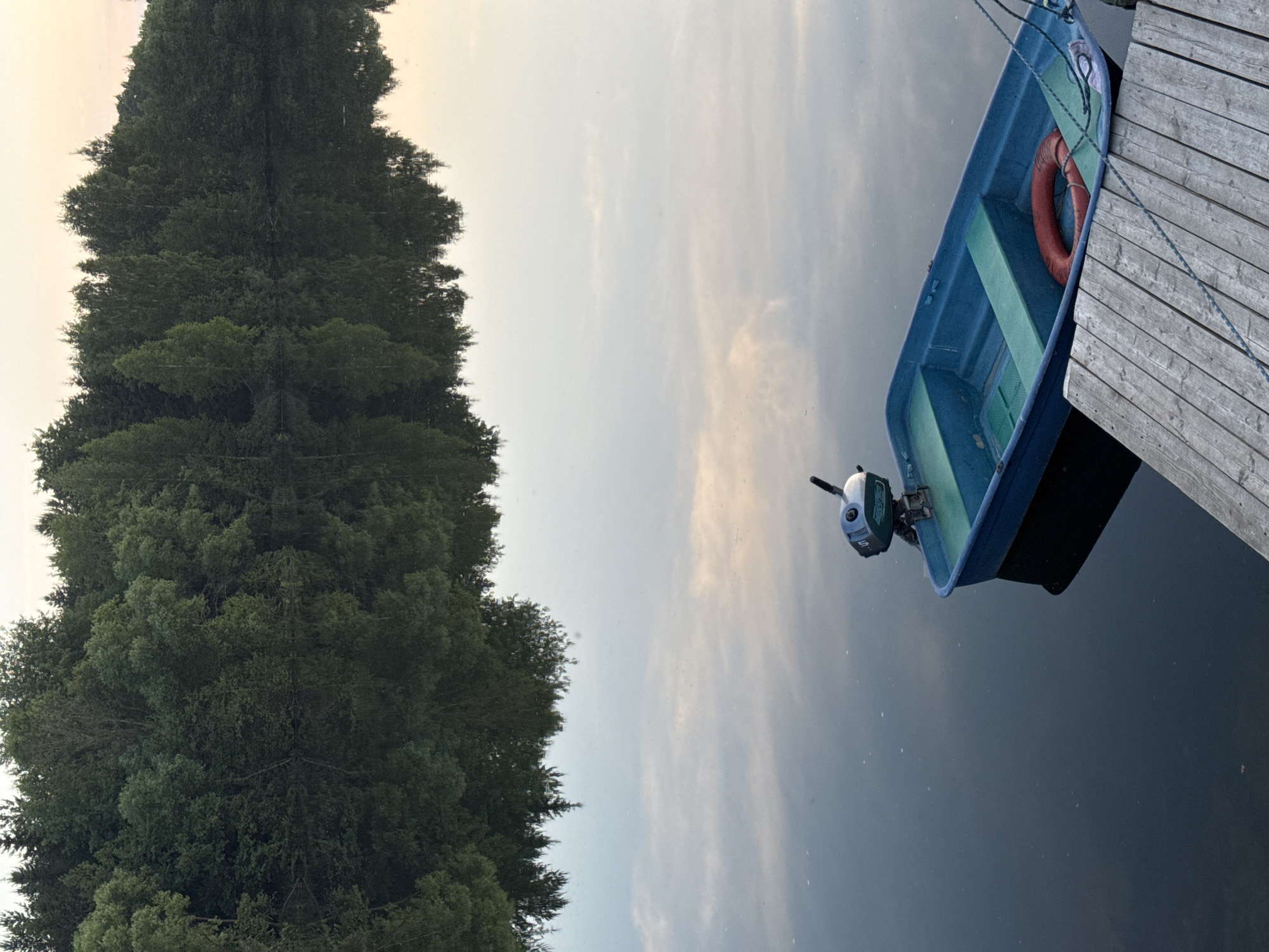 A blue boat with a small outboard motor is docked at a wooden pier on a calm body of water. The boat is empty and contains a red life preserver. The opposite shore is lined with a dense forest of green trees, whose reflections are visible on the water's surface. The sky above is a pale, muted color, suggesting either dawn or dusk. The image appears to have been taken in Kamysh, Russia.