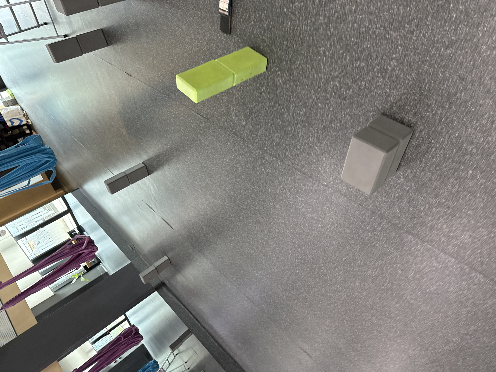 The image shows a yoga studio with aerial yoga hammocks hanging from the ceiling. There are several yoga blocks scattered on the grey floor, some stacked on top of each other. A bright green yoga block is prominently placed in the foreground, leaning against another block. In the background, there are large windows and a mirror, reflecting more of the studio space. The overall lighting is bright, suggesting an indoor daytime scene. There are no people visible in the image.
