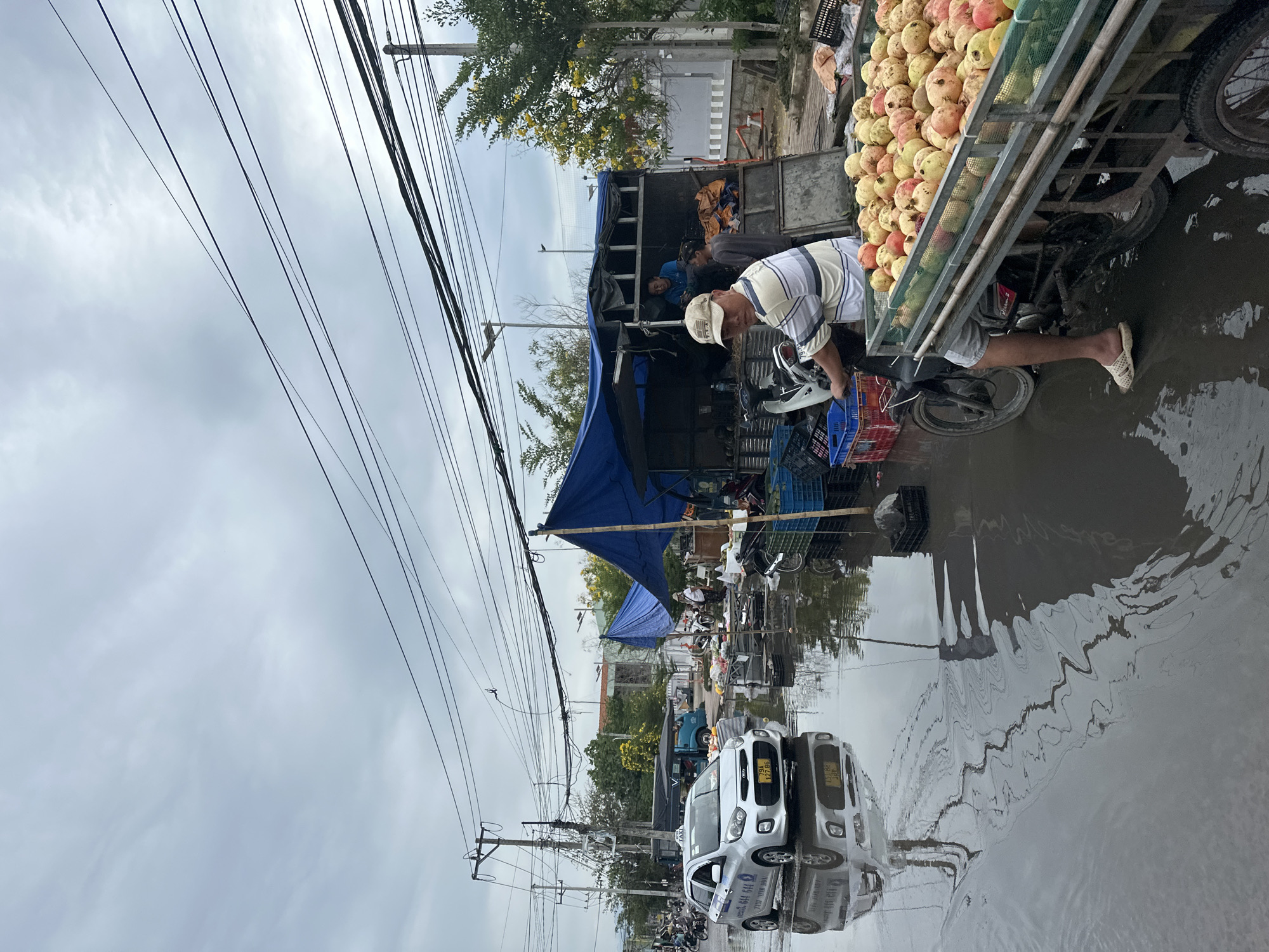 The image depicts a flooded street in Nha Trang, Vietnam, likely after heavy rainfall. A silver Kia Morning taxi is driving through the water, its reflection visible on the wet surface. On the right side of the street, a makeshift market is set up under blue tarps. A man wearing a striped shirt and a cap is operating a motorcycle with a trailer loaded with pomegranates. He is positioned in the foreground. Behind him, two other men are visible near a truck. The scene suggests a typical street market in Vietnam, adapted to cope with the flooded conditions. Electrical wires crisscross the sky, typical of urban environments in Vietnam.