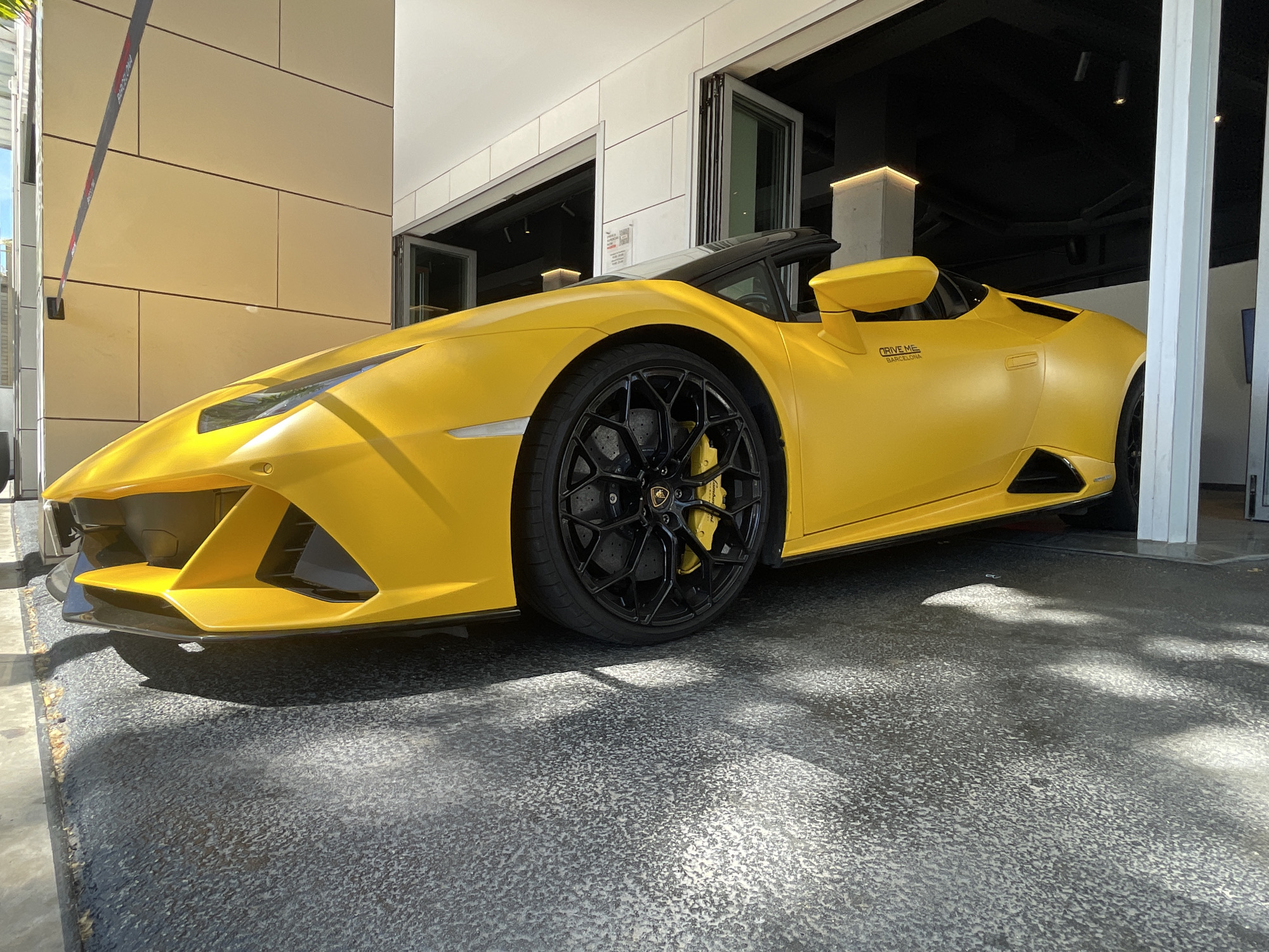 A vibrant yellow Lamborghini Huracán Spyder, appearing to have a matte or satin finish, is prominently displayed, partially indoors and partially outdoors, in Barcelona, Spain. The sleek, convertible sports car is facing towards the left of the frame. It features black multi-spoke wheels with yellow brake calipers visible through the spokes, matching the car's body color. A sticker on the side of the car, near the rear wheel arch, clearly reads "DRIVE ME BARCELONA," confirming the location.
The car is parked on a dark gray, textured ground, possibly asphalt or concrete, with distinct shadows cast by the bright natural sunlight, indicating it is daytime. The background consists of a modern building with light-colored, possibly beige, rectangular wall panels on the left. On the right, large glass folding doors lead into an interior space characterized by a dark, possibly black, ceiling with modern track lighting. A white pillar is also visible within the indoor section. A red and white barrier with "BARCELONA" printed on it can be seen partially in the upper left background, further cementing the location context. No people are visible in the image.