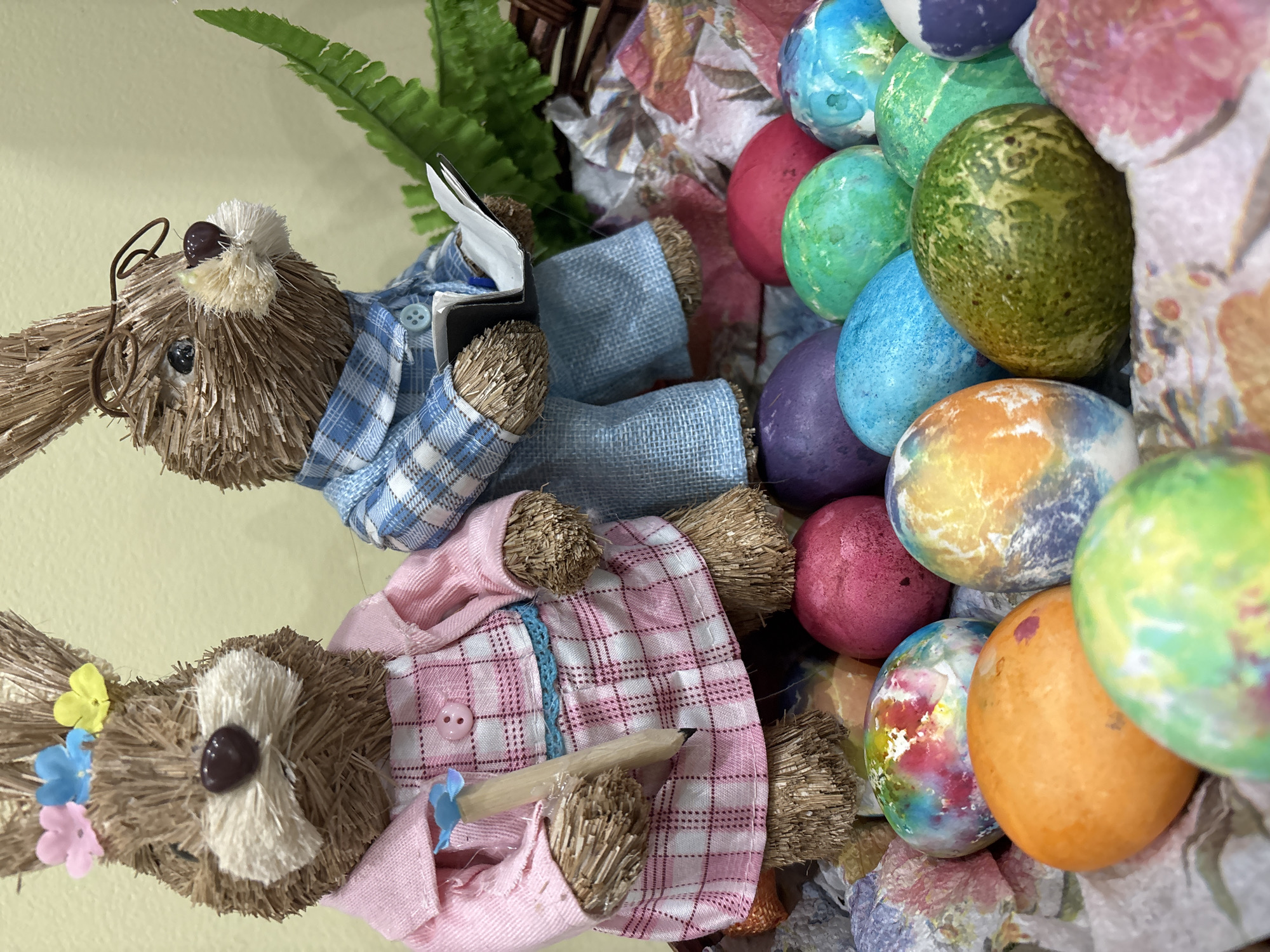 The image depicts a wreath with two straw bunny figures and numerous colorful Easter eggs. One bunny is wearing a blue plaid shirt and eyeglasses, holding a notebook, while the other is wearing a pink plaid shirt and holding a pencil. The eggs are painted in various colors and patterns, including tie-dye effects. The wreath is made of straw and includes artificial greenery and floral decorations. The background is a plain pale yellow. There is no indication of the location being in Sofia, Bulgaria, or any specific city.
