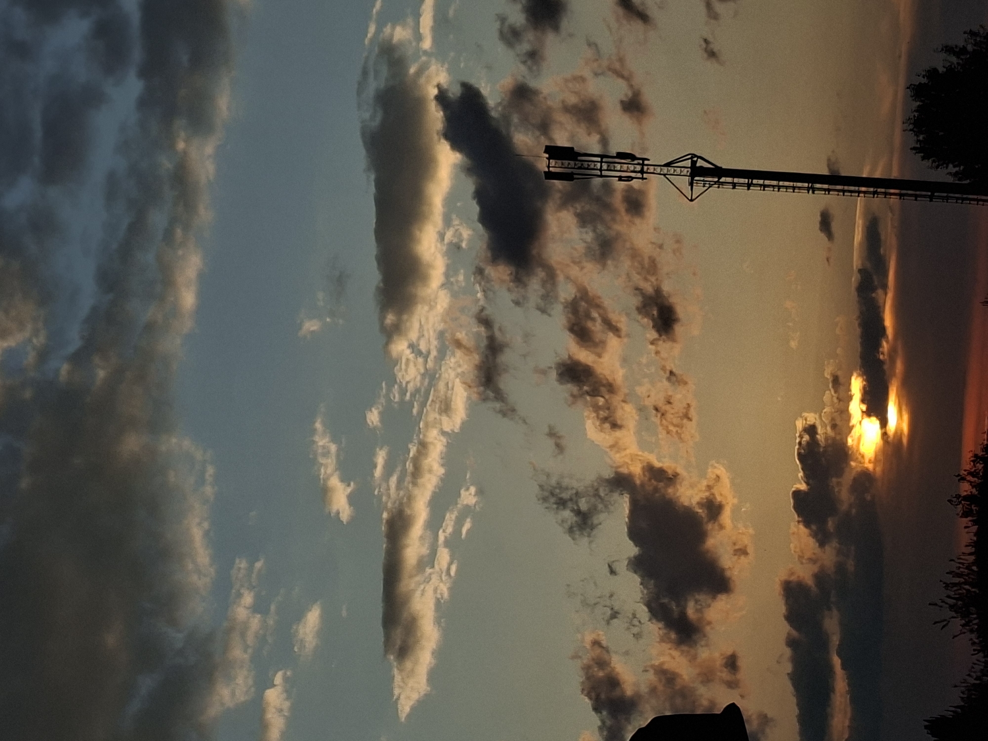 The image shows a view of the sky at sunset. The sun is low on the horizon, appearing as a bright light shining through the clouds. Clouds are scattered throughout the sky, with varying shades of gray and white. The sky ranges in color from blue to orange. A utility pole is visible, stretching horizontally across the frame, its silhouette outlined against the colorful sky. Tree silhouettes can be seen on the right side. Location cannot be determined from the given image.