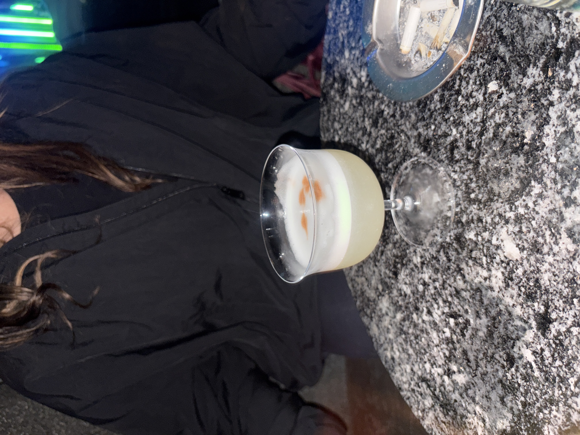 The image shows a bar scene. A person wearing a black jacket sits at a table with a grey, speckled surface. A cocktail, likely a Pisco Sour due to its frothy white top and reddish-brown bitters design, sits on the table. An ashtray with cigarette butts is also present. The location is possibly in Buenos Aires, Argentina, as Pisco Sour cocktails are popular in South America. The scene depicts a casual, possibly late-night setting.