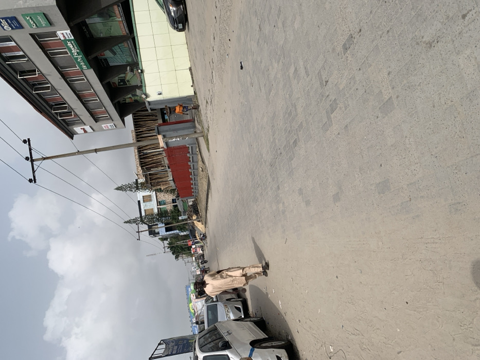 The image depicts a street scene in Jakande, Nigeria, under an overcast sky. The foreground shows a sandy shoulder transitioning into a grey interlocking paver road surface. On the left, a man wearing a light-colored tunic and trousers walks away from the camera, holding a dark object in his right hand, past parked white and silver vehicles. On the right, a multi-story building with a light-colored tiled base and red-framed windows displays business signs including "MILAN" and "Schneider Electric." A pedestrian in an orange top and dark shorts walks away from the camera along the building's base, carrying an item on their head. Further down the street, a red corrugated fence fronts a building under construction with wooden scaffolding, and utility poles with overhead power lines line the street. A black sedan is partially visible parked near the multi-story building on the far right.
