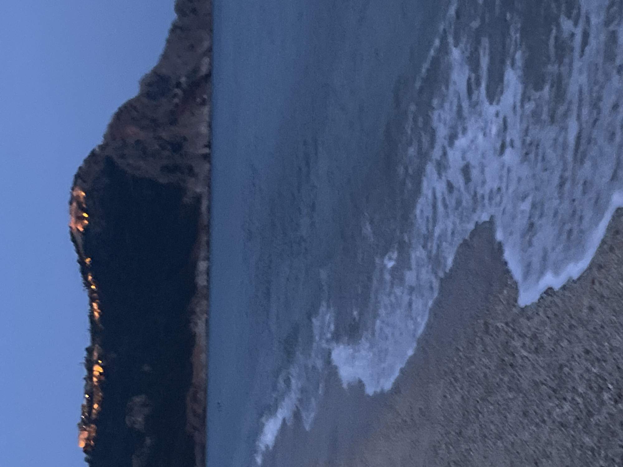 The image depicts a beach scene in Alanya, Turkey, at dusk or dawn. A dark, rocky cliff borders the left edge of the frame, with faint glimmers of light suggesting possible structures or lights along its face. Adjacent to the cliff, the sea transitions from a deep blue color further away from the shore to a lighter, foamy white as the waves break onto the sandy beach in the lower right quadrant of the image. The beach itself is a coarse, dark sand. No people or identifiable objects are visible.