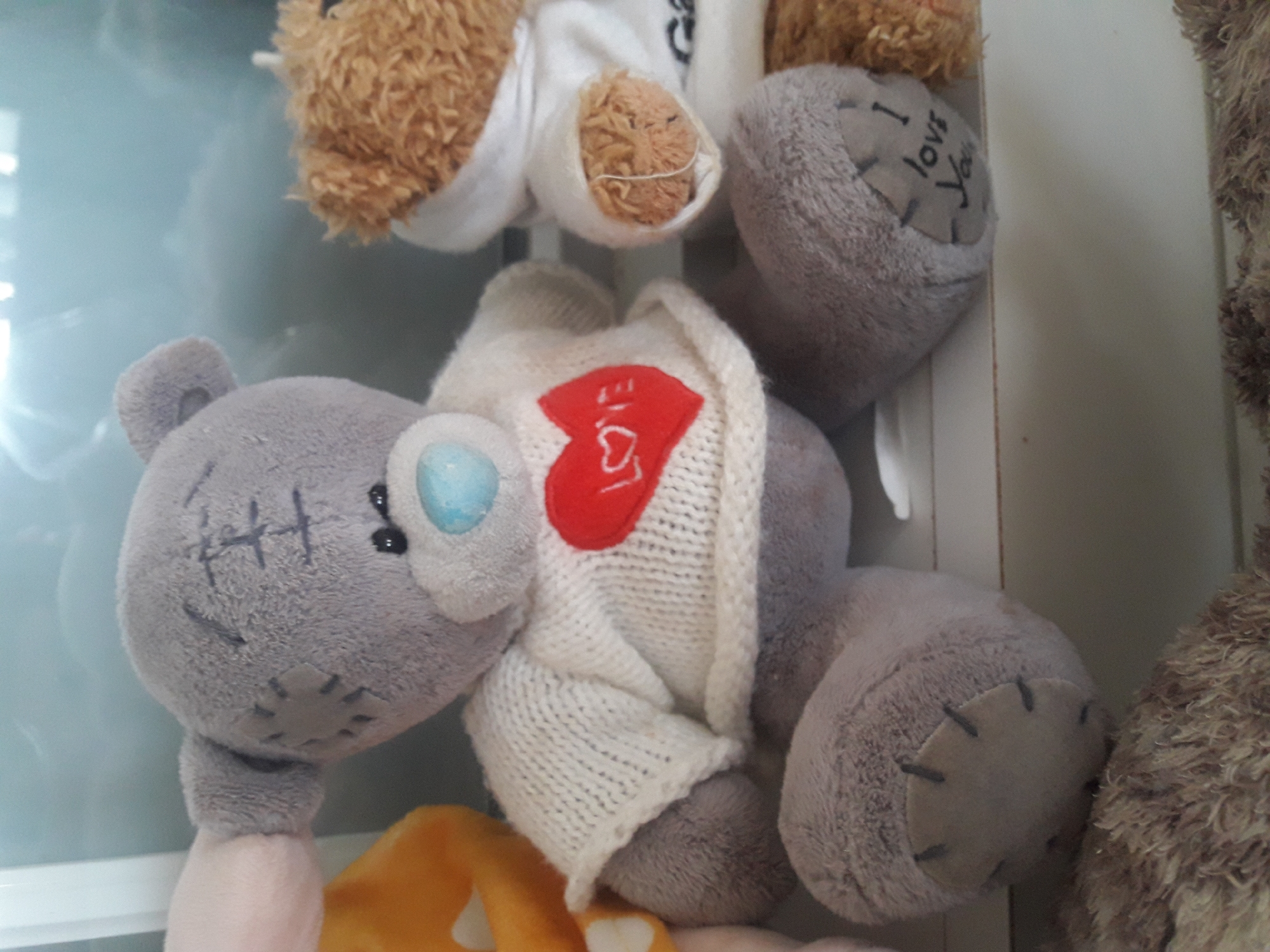In an indoor setting, three teddy bears are displayed on a white shelf. The leftmost bear is a light pink color and is partially visible. In the center, a grey teddy bear with a white knitted sweater and a red heart with the word "Love" embroidered on it is positioned. The third teddy bear, to the right, is brown with a white hoodie and has the words "I love You" written on its left paw. The background appears to be a reflective surface, possibly a window or glass, showing a blurred reflection of an interior space with some light sources. The overall scene is static, with no observable actions or interactions. The location is unknown.