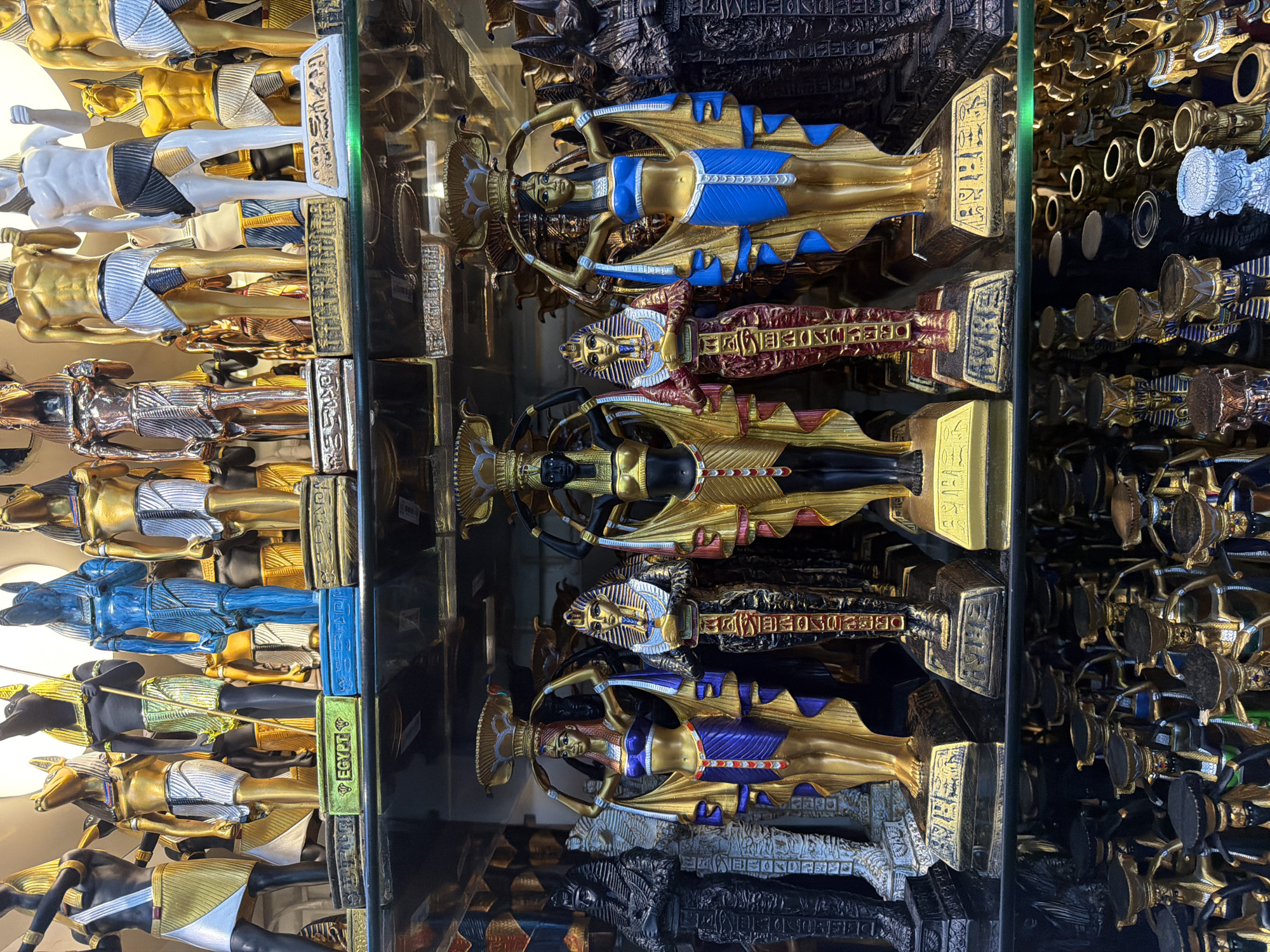 The image shows a retail display of Egyptian-themed figurines, likely souvenirs, in Sharm El Sheikh, Egypt. The figurines are primarily gold and black, with some in blue and red accents. They depict pharaohs, queens, and deities such as Anubis and a blue-skinned god. The figures are arranged on shelves, with several rows visible. The top shelf features taller, more prominent figurines, while the lower shelves display smaller, more numerous items. The environment appears to be an indoor market or shop, with artificial lighting. There are no people visible in the image. The overall impression is one of abundance and variety of Egyptian cultural artifacts for sale.