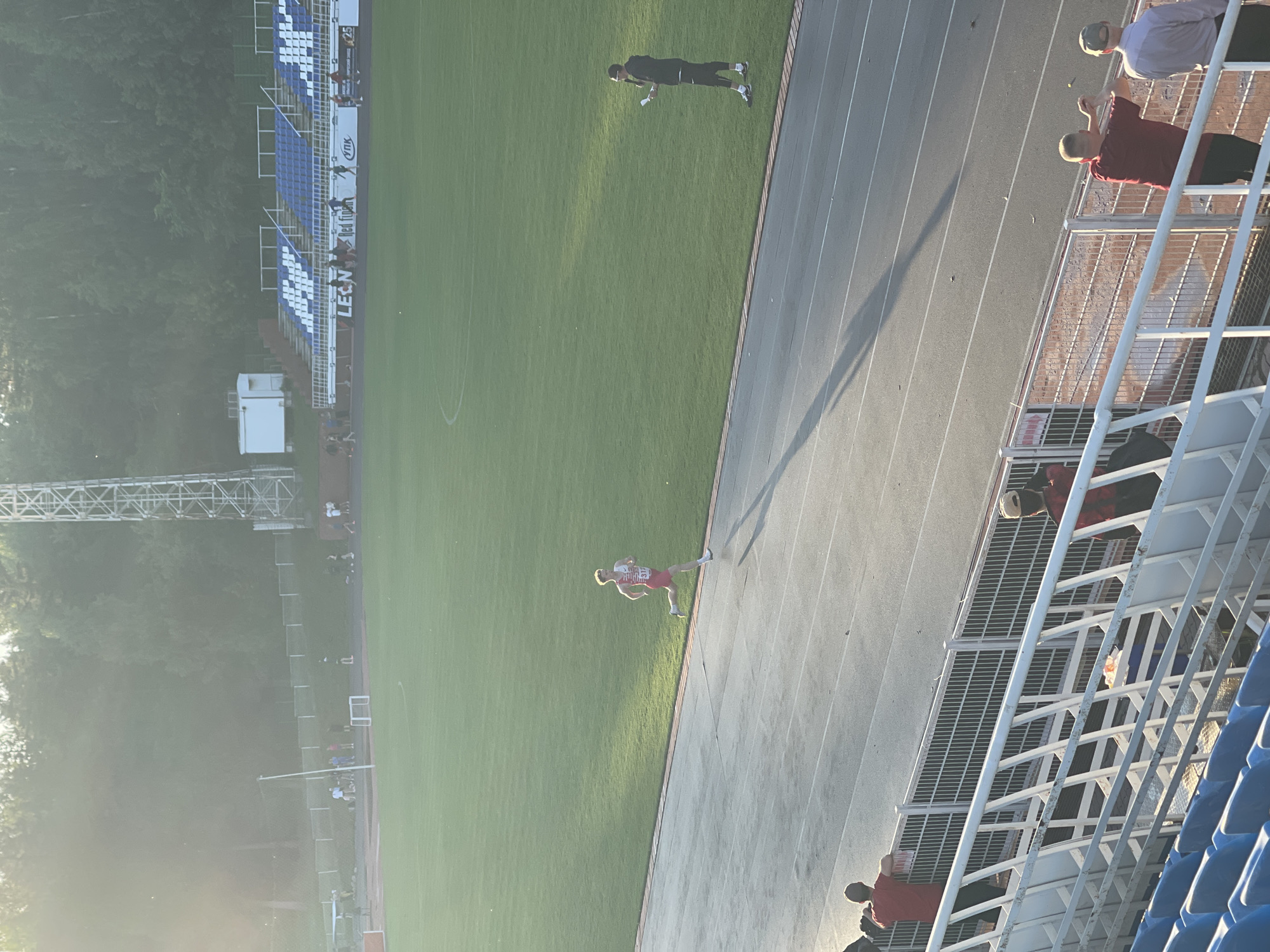 An outdoor athletic track and field event is taking place. In the foreground, several spectators are visible sitting and standing in bleachers, with a metal fence separating them from the track. The track itself is a dark grey surface with white lines marking lanes. In the middle ground, a male runner in a red and white uniform is sprinting on the track, casting a long shadow. To the right of the track, on the grass field, a male individual wearing glasses and holding papers stands and appears to be addressing the runner or observing the event. In the background, a large green field, stands with blue and white seats, trees, and a tall metal tower are visible. The sun is low in the sky, creating a bright lens flare effect and casting long shadows across the scene.