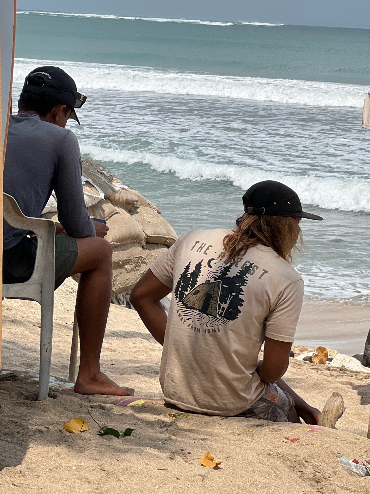 Two individuals are seated on a sandy beach in Kuta, Indonesia, facing the ocean. The individual on the left, wearing a dark long-sleeved shirt and dark shorts, sits on a white plastic chair and appears to be holding a phone. The individual on the right, wearing a tan t-shirt with a graphic print and patterned shorts, is seated on the sand, looking out at the waves. The t-shirt graphic depicts a campsite with a tent and trees. The ocean is rough with visible waves. Behind the seated individuals, sandbags are stacked, and debris is scattered on the sand.