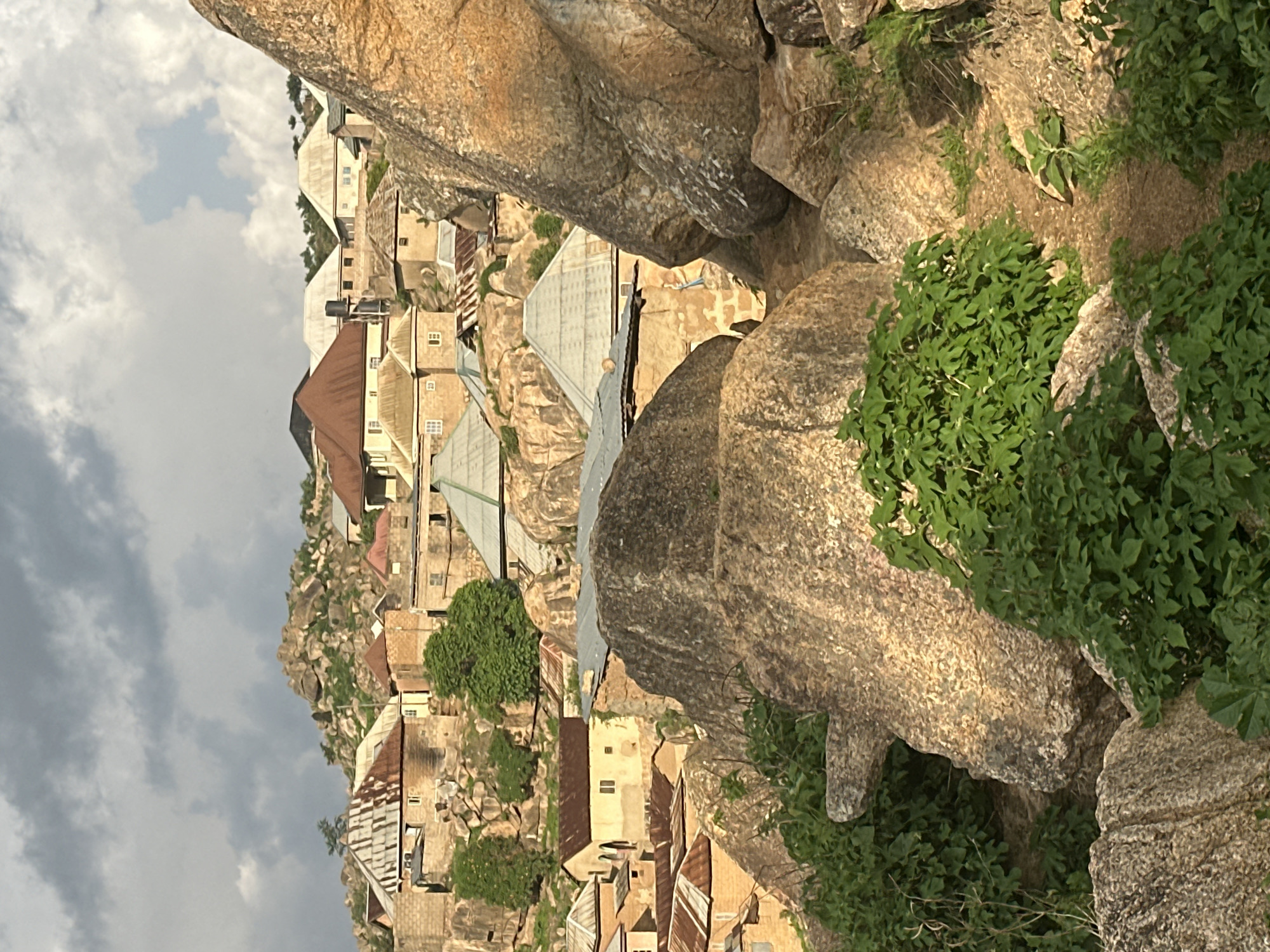 This image depicts a dense residential settlement intricately integrated with massive granite rock formations, characteristic of the Jos Plateau in Nigeria. The scene is dominated by numerous single-story and some two-story houses built directly into and around the large, weathered boulders.
The houses feature light brown or beige walls, likely constructed from stone or concrete blocks, and are topped with a mix of corrugated metal roofs. Some roofs exhibit a rusted, reddish-brown hue, while others are newer and silvery-grey. Small, rectangular windows are visible on the building facades, suggesting a practical and established architecture. The dwellings are closely clustered, indicating efficient use of the challenging terrain.
Prominent, colossal granite boulders are a defining feature, not only framing the view but also serving as natural foundations, walls, and boundaries for the houses. Green vegetation, including dense bushes and smaller plants, grows abundantly in the crevices of the rocks and around the buildings, providing a natural contrast to the stone and man-made structures. A few small trees are also scattered throughout the landscape.
The sky is partly cloudy, with significant grey clouds covering parts of the frame, suggesting either an overcast day or a specific time like early morning or late afternoon. The lighting is soft, casting some areas into shadow while others are gently illuminated. No human activity or visible text is discernable. The image captures a serene yet resilient community where architecture and nature coexist in a striking symbiotic relationship within the unique landscape of Jos, Nigeria.