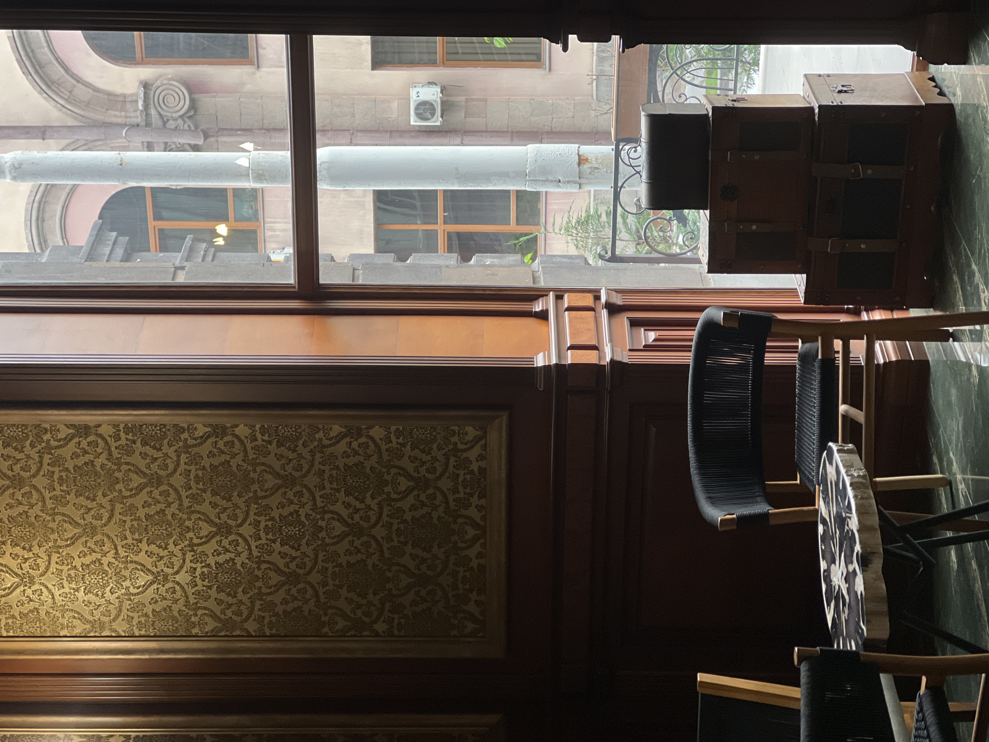 The image, potentially taken in Yerevan, Armenia, shows a section of an interior space with a view of an exterior scene. A window occupies the upper portion of the frame, revealing an exterior facade of a building with architectural details, including rounded window frames, ornate designs, and what appears to be a utility pipe running horizontally across the frame. Additionally, an air conditioning unit is visible on the exterior wall.
Inside, the image features ornate wooden paneling with a decorative, textured wallpaper or fabric. Two black woven chairs are partially visible, positioned near a wooden table. To the right of the window, luggage trunks are stacked. The lighting is dim, suggesting an intimate or subdued atmosphere.