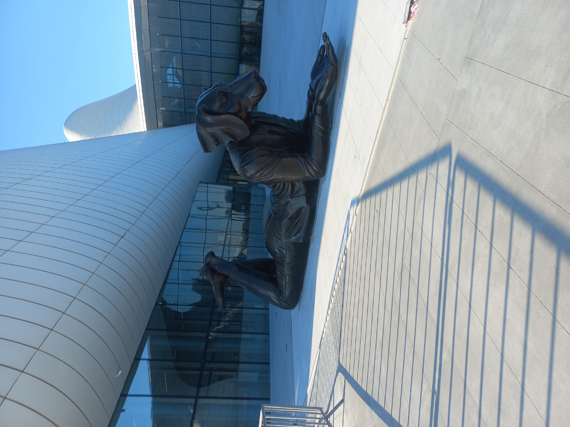 This image captures a striking sculpture of a bronze dog lying down, positioned outside the Heydar Aliyev Center in Baku, Azerbaijan. The sculpture is in the foreground, with its legs bent and extended behind it, and its arms outstretched, with hands open. The dog is wearing a suit, indicating a human-like anthropomorphism. Behind the sculpture, the iconic, fluid architecture of the Heydar Aliyev Center is visible, characterized by its white, curved facade and large glass windows reflecting the clear blue sky. The ground in front of the sculpture is paved with large, light-colored square tiles, and the shadow of a metal fence is cast across them, creating a pattern of parallel lines. The lighting suggests it is daytime, with the sun casting strong shadows. There are no people visible in the frame.