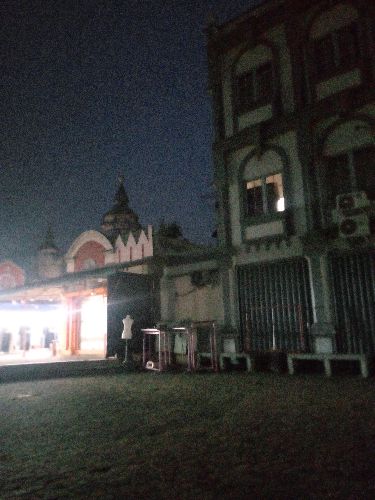The image shows a building with ornate architecture, featuring arched windows and decorative elements. To the left, a structure with multiple domes, possibly a religious building, is visible. In the foreground, the ground appears to be paved and somewhat worn. Several objects are present, including what looks like a mannequin, and various stands or racks, suggesting a retail or exhibition space. The scene is lit by artificial lights, indicating it is nighttime. The overall impression is that of an exterior view of a commercial or public area in a city at night. The specific location identified is Cikupa, Indonesia.