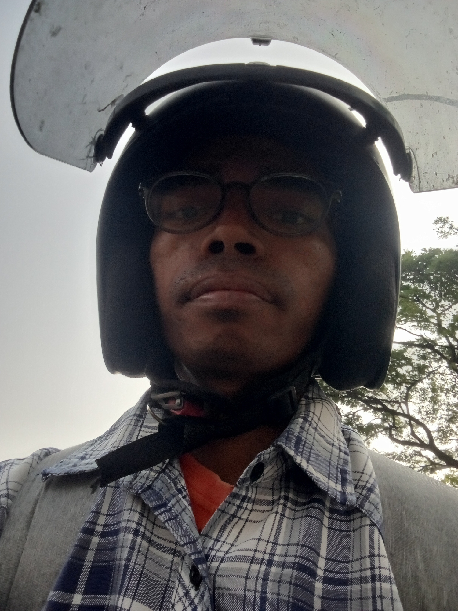 A person wearing a black motorcycle helmet with a clear visor is pictured in a close-up, self-taken shot. The helmet obscures the top of their head and forehead, but their eyes are visible behind round, dark-rimmed glasses. They have a mustache and stubble. They are wearing a blue and white plaid shirt over an orange shirt, and a gray jacket. The background shows a slightly overcast sky and the branches of a large tree, suggestive of an outdoor setting in Surabaya, Indonesia.