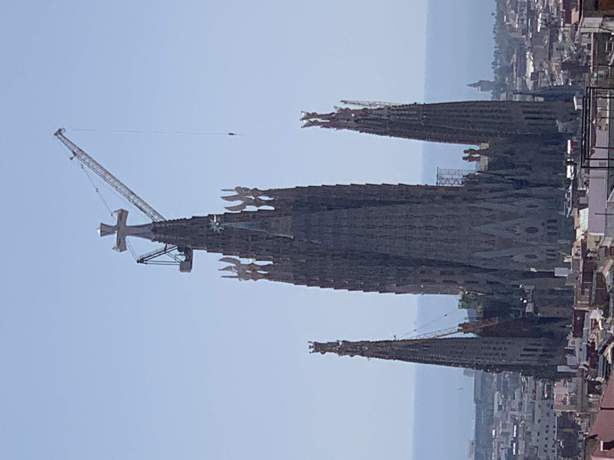 This is an aerial, eye-level, medium shot of the Sagrada Familia basilica in Barcelona, Spain, on a clear, sunny day. The basilica is a large, gothic-style structure made of dark stone, with multiple spires. The central spire is the tallest, topped with a large cross. A construction crane is attached to the central spire, with its arm extended to the right. A thin cable hangs from the crane. Several smaller spires surround the central spire. Below the basilica, the cityscape of Barcelona is visible, with many buildings of varying heights and colors, and a hint of the sea in the background. The sky is a pale blue.