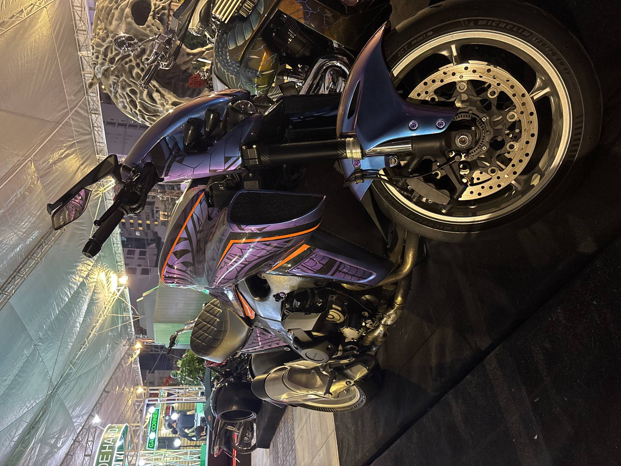The image captures a custom motorcycle with intricate paintwork and a skull-themed decorative element, possibly at an event in or near Nha Trang Ward, Vietnam. The motorcycle features a metallic purple and blue color scheme with geometric patterns, an aftermarket exhaust, and high-performance Michelin tires. Additional motorcycles and decorative elements, including a large skull, are visible in the background. The setting appears to be an event space, possibly under a tent structure, with other exhibitors or displays in the far background. The lighting suggests an indoor or nighttime environment.