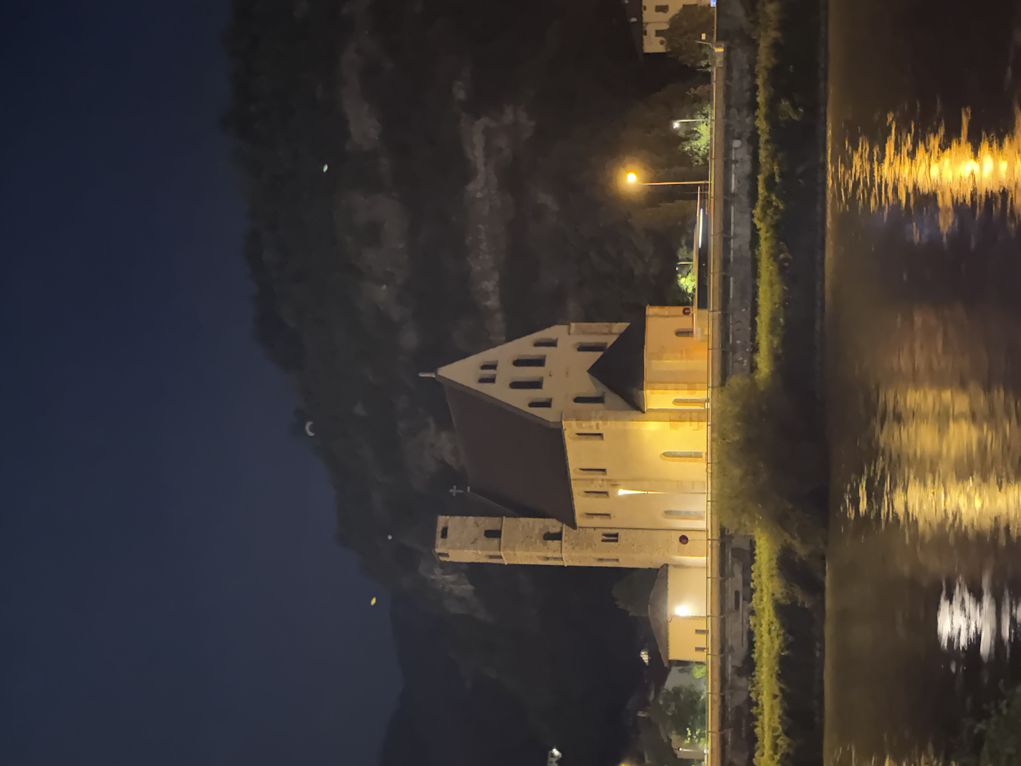 A church with a tall bell tower is illuminated at night, situated on a hill overlooking a river in Trento, Italy. The church's facade is a light color, likely stone, and features a cross at the apex of its roof. The bell tower is a similar stone construction, with visible windows. The river in the foreground reflects the lights from the church and surrounding streetlights. A lush green embankment separates the river from the man-made structures. Beyond the church, a dark, tree-covered hill rises into the night sky. Streetlights line a road behind the church, and a blurred vehicle is visible on this road, indicating movement. Several other buildings, some also lit, are visible in the background.