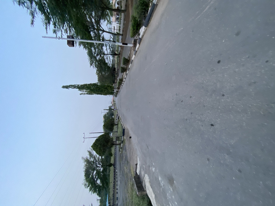 An empty, straight asphalt road extends into the distance under a pale blue sky in Osu, Nigeria. The grey road surface shows minor irregularities and scattered dark spots. On the left, a black and white painted curb borders a wide, unpaved area. This area contains a large, dark paved section with numerous black and white concrete blocks, alongside various trees, utility poles with overhead power lines, and a partial view of a blue-roofed structure in the far background. A small, dark, unidentifiable object rests on the paved ground near the left curb. On the right, a similar black and white curb outlines a landscaped strip featuring a row of tall, slender evergreen trees and other foliage. A light pole with a dark, rectangular fixture is prominent on the right, and light-colored buildings with archways are visible in the far-right background. No people or vehicles are present.