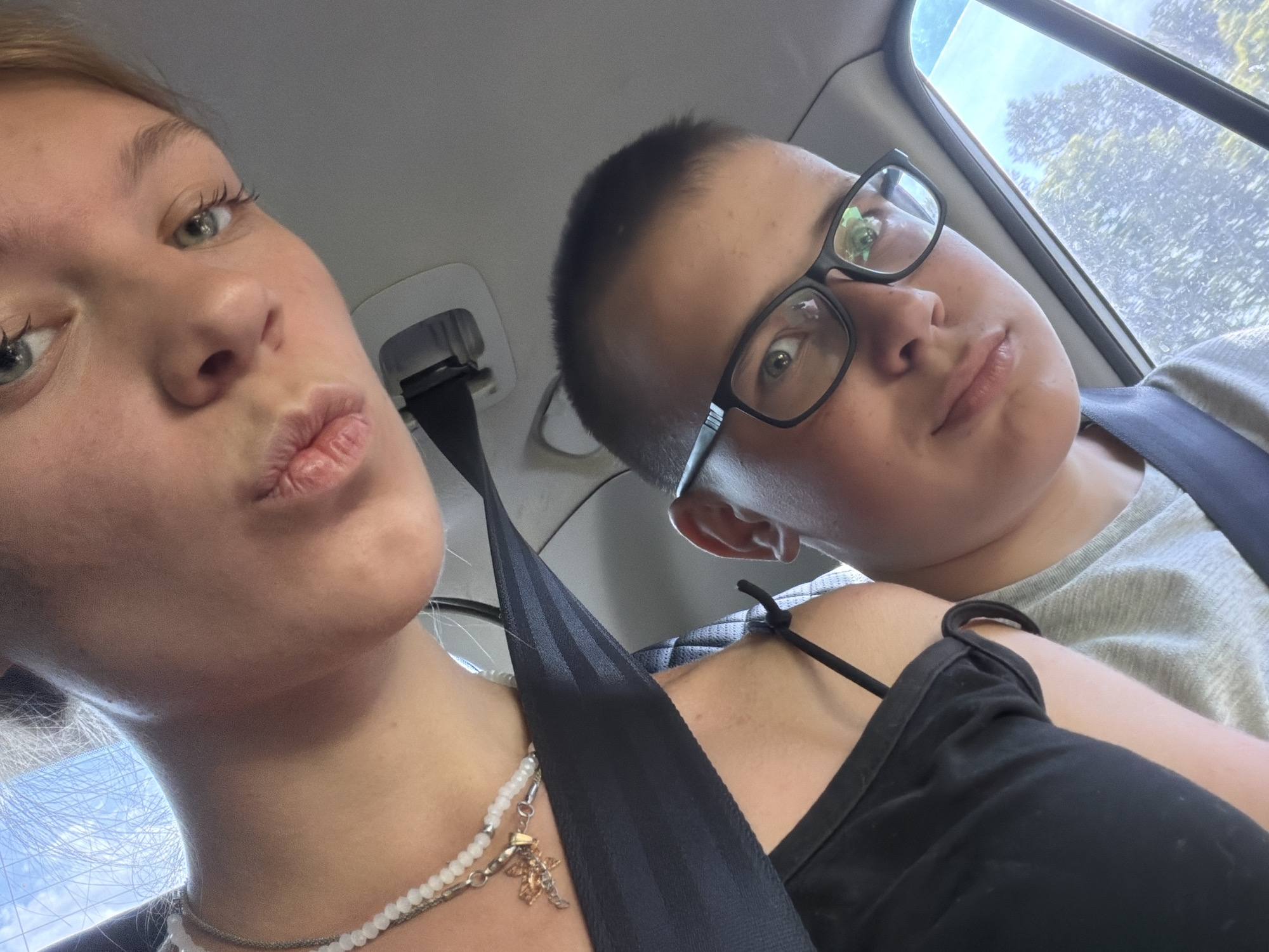 In Kruglovka, Russia, two individuals are visible within a vehicle, captured in a selfie. The person in the foreground, positioned on the right, is a young male wearing glasses and a grey t-shirt, with a seatbelt visible across his chest. He is looking towards the camera. Behind him, to the left, a young female is also visible, with her face angled downwards and her lips pursed. She is wearing a black top and a beaded necklace with a pendant. Her seatbelt is also visible, crossing her shoulder. The interior of the car, including part of the headliner and a car door, is discernible. The background through the car window suggests an outdoor environment with trees and foliage, indicative of a possibly rural or suburban setting outside of Kruglovka. The lighting suggests it is daytime.