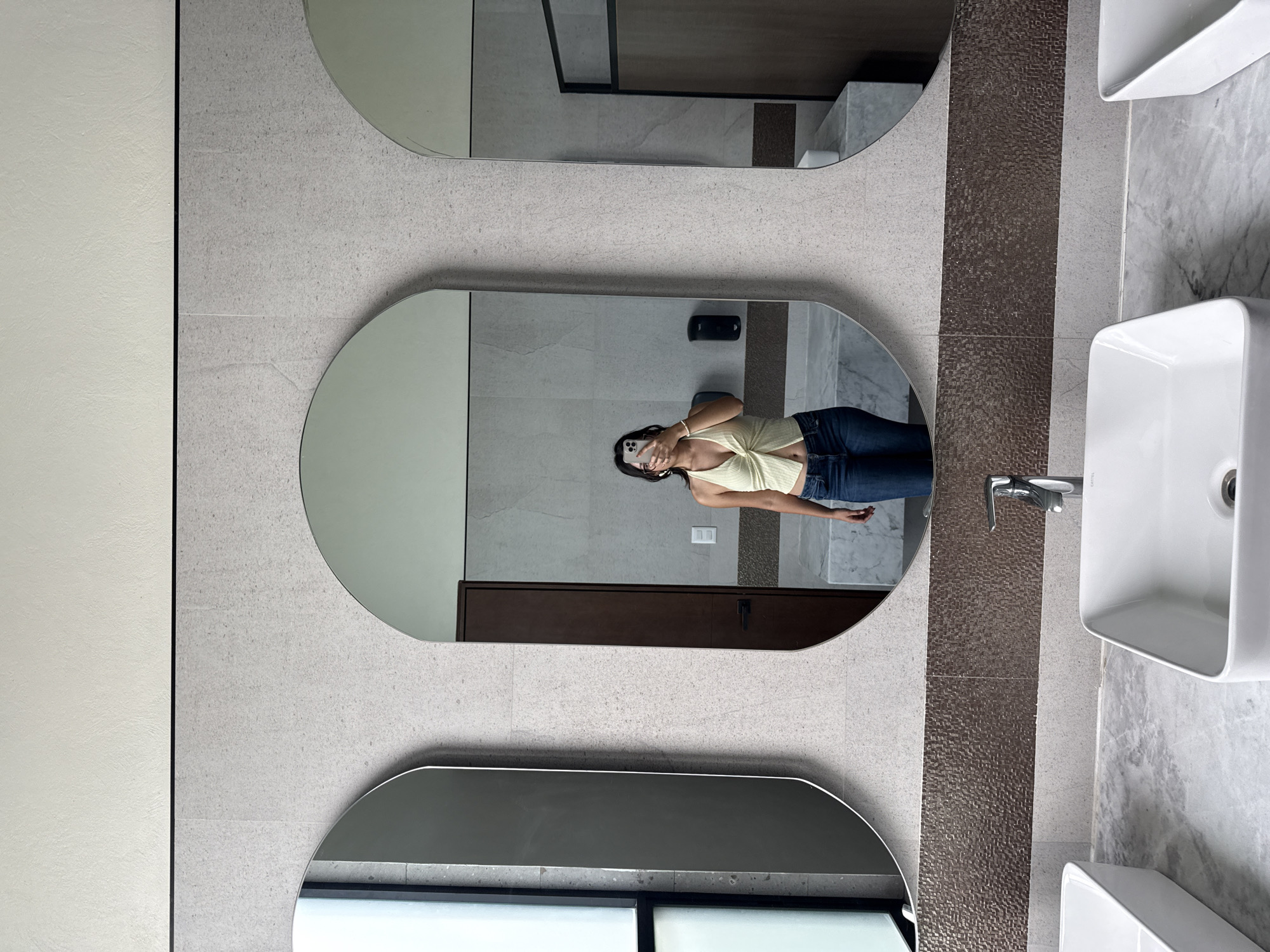 A woman is reflected in a bathroom mirror. She is wearing a light-colored tank top and blue jeans, holding a cell phone. The bathroom has three mirrors and two white square sinks. The mirrors and sinks are mounted on a wall with gray tile. There is a strip of brown mosaic tile separating the mirrors from the sinks. The location is Santa Ana Yancuitlalpan (Chilhuacán), Mexico.