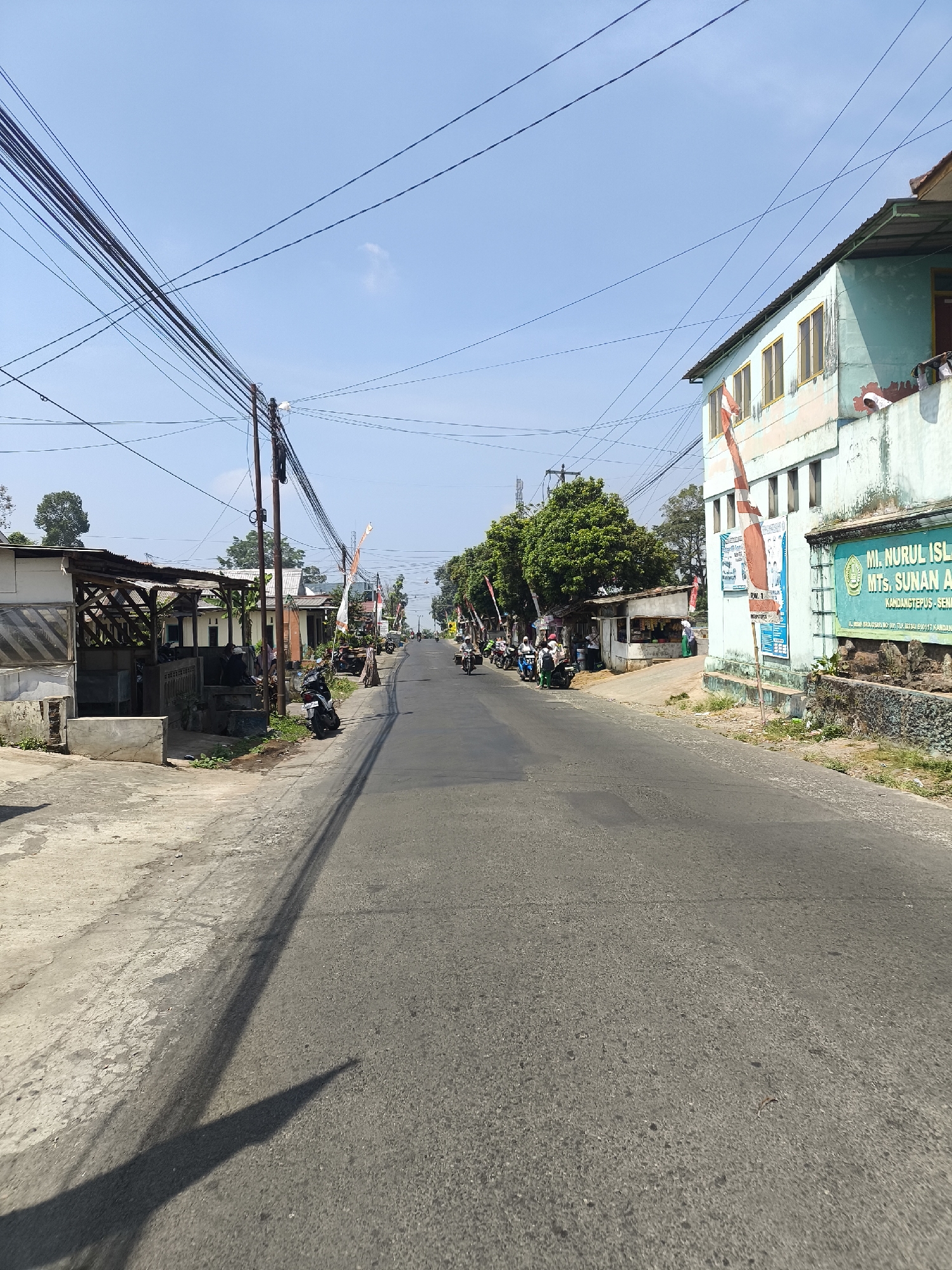 The image depicts a street scene in Senduro, Indonesia, on a clear, sunny day. A paved road runs through the center of the frame, leading into the distance. On the right side of the road, a light blue building with a green sign that reads "MI. NURUL ISLAM MTS. SUNAN AM...", and other text in Indonesian, is visible. Red and white Indonesian flags are attached to the building and a pole in front of it. Several trees with green foliage line the right side of the road. On the left side of the road, there are several structures with open-air coverings and what appear to be small businesses or stalls. A motorcycle is parked on the left side of the road in the foreground. Further down the road, several motorcycles are in motion, with people riding them. Utility poles with numerous wires stretch across the sky. The overall environment suggests a rural or suburban setting within a town or village.