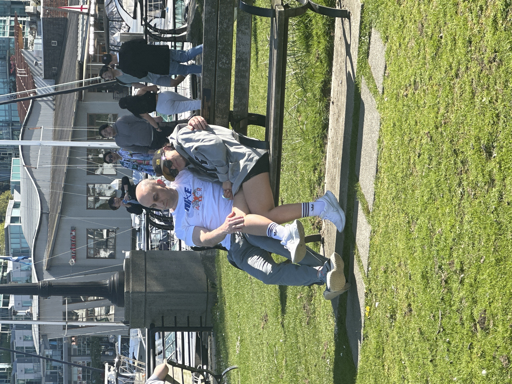 A bald man wearing a white Nike shirt and grey pants sits on a park bench in Vancouver, British Columbia. A woman with long hair wearing a grey hoodie and black shorts is lying on the bench with her head on the man's lap. They are located near the water with boats in the background. The location is likely near Granville Island as a sign for "TAPERO'S" can be seen on a building in the background. The weather is sunny. There are people walking in the background.