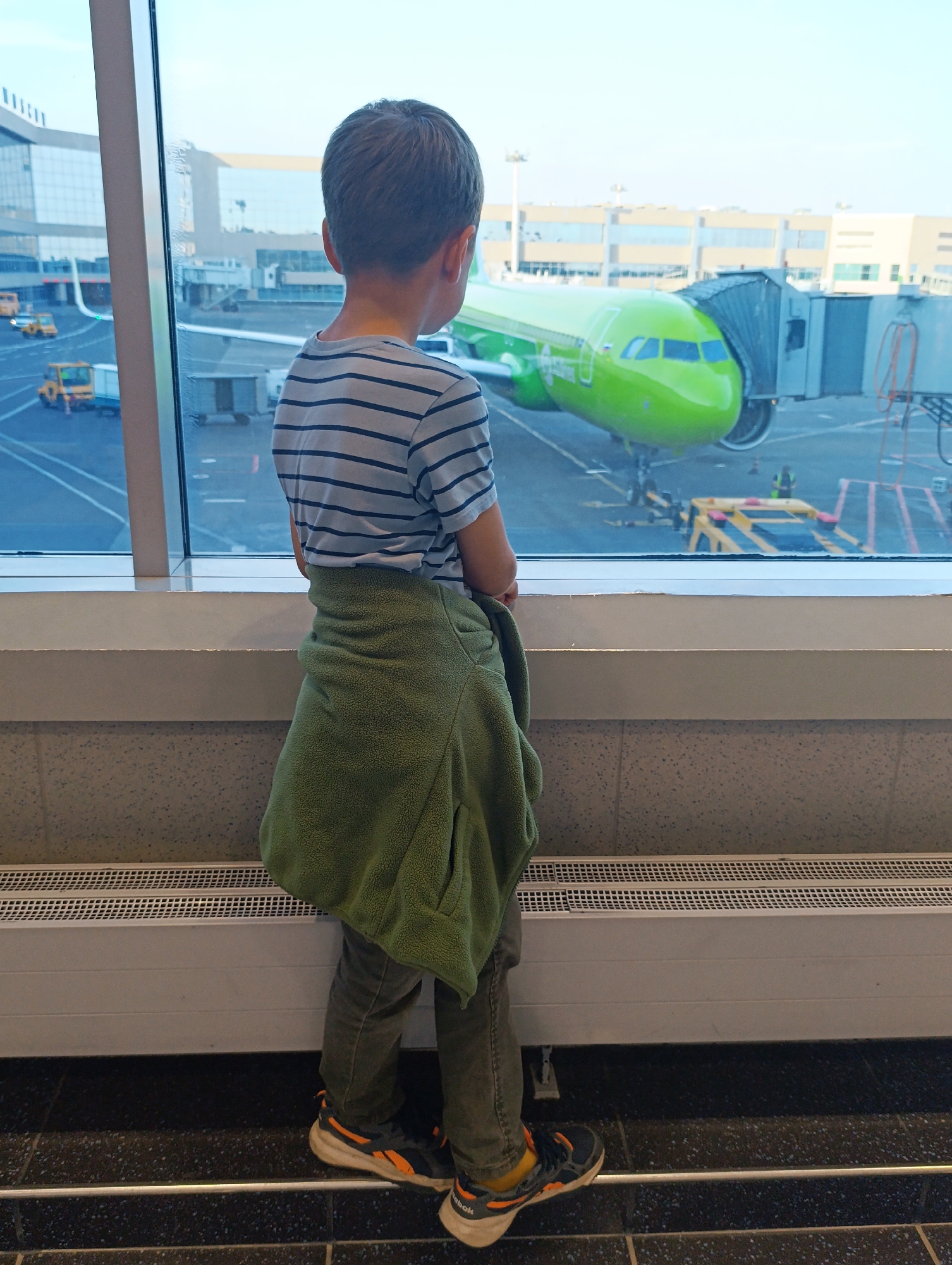 At Domodedovo International Airport in Domodedovo, Russia, a young boy stands with his back to the camera, looking out a large window at a bright green airplane parked at a jet bridge. The boy wears a striped t-shirt and green fleece jacket draped over his shoulders. He is also wearing dark pants and athletic shoes. The airport terminal building is visible to the left, with several yellow airport vehicles on the tarmac. The sky is bright and clear.