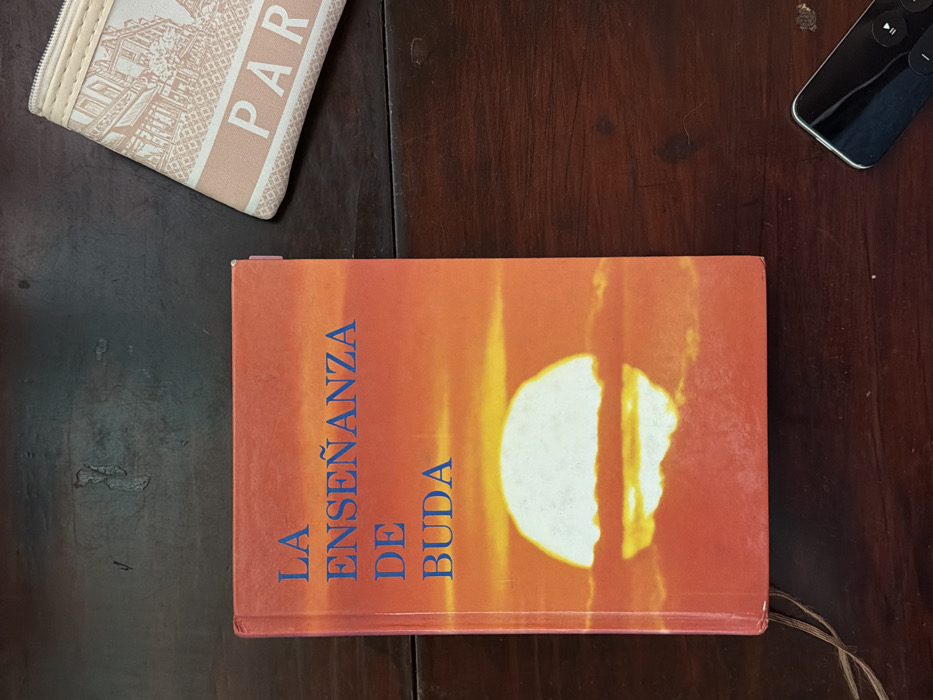A close-up shot presents a book titled "LA ENSEÑANZA DE BUDA" on a dark wooden surface. The book's cover features a sunset-like image. In the upper left corner, part of a beige wallet with "PARIS" printed on it is visible. A black remote control sits on the right side of the frame, also on the wooden surface. The scene conveys a sense of reading or relaxation.