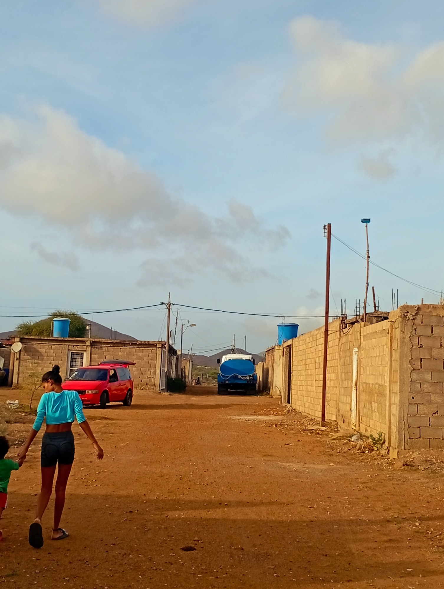 The image depicts a dusty, unpaved street in what appears to be a residential area. In the foreground, a woman with dark hair pulled back in a bun walks away from the camera, holding the hand of a child wearing a green shirt and red shorts. The woman is wearing a bright blue cropped top and dark shorts. Further down the street, a red vehicle is parked. To the right of the street, buildings constructed from cinder blocks are visible. Several large blue water tanks are present, one on top of a building and another on a truck. A utility pole with wires runs along the street. The sky is a light blue with scattered clouds. The scene suggests a rural or semi-urban environment.