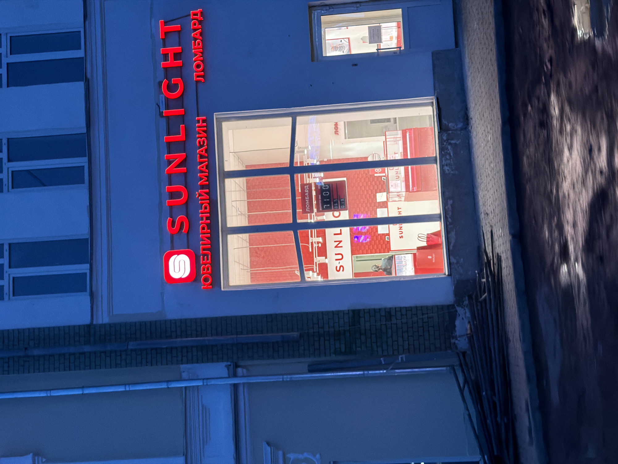 Here is a forensic description of the image:
The image shows the exterior of a "SUNLIGHT" jewelry store and pawnshop ("ЛOMБAPД") located in what appears to be a residential area in the city of Sarapul, Russia. The storefront has a large window with a red interior and the store's sign is illuminated. A smaller window above the storefront also appears to be lit. The street and possibly a sidewalk are visible in the foreground. The building's facade is blue and white, and a row of windows from another part of the building can be seen to the left of the store.