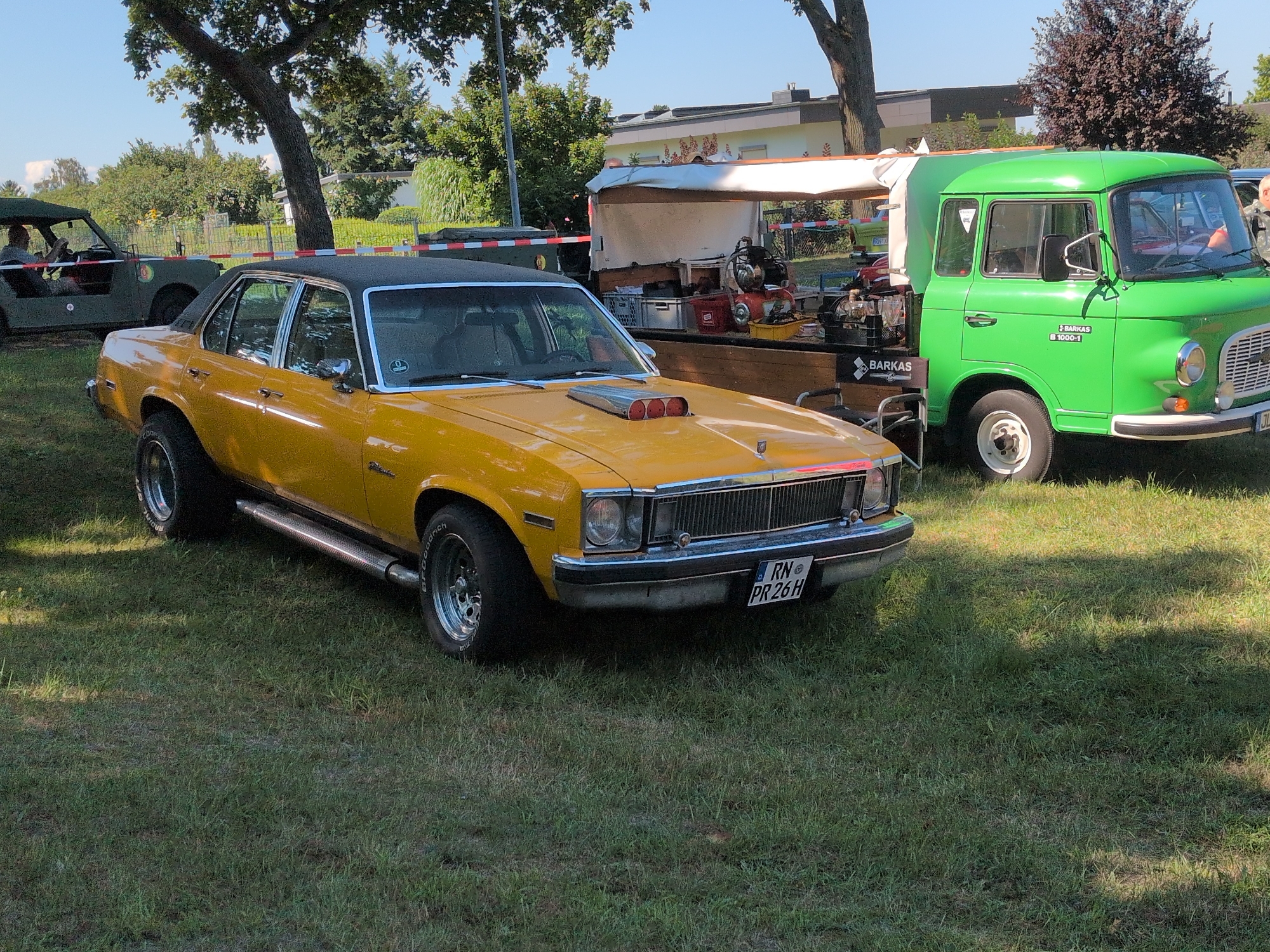 In Hohenferchesar, Germany, a yellow 1970s era American muscle car with a black vinyl roof and chrome accents is parked on a grassy area. The car has a prominent hood scoop with three air intakes. To the right of the muscle car is a bright green Barkas B 1000-1 van, also parked on the grass, with its side door open. Behind the Barkas van, a white canopy covers a stand that appears to be selling drinks or food, with various items and equipment visible. In the background, trees and buildings are present, suggesting a rural or suburban setting. The sky is clear and blue. The ground is covered in green grass.
