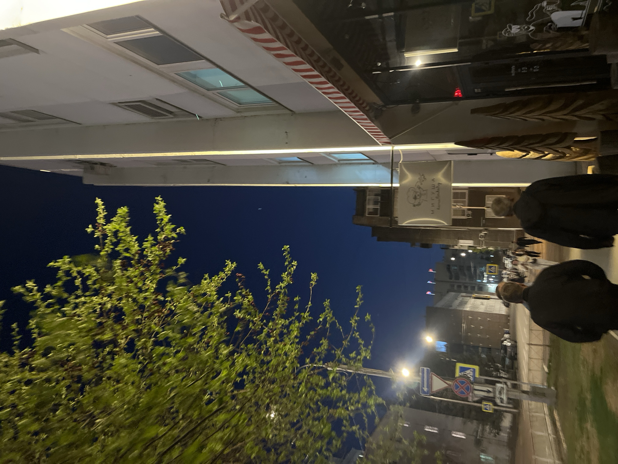 The image depicts a street scene in Krasnoyarsk, Russia, at night. A building with skylights and a striped awning overhangs the sidewalk. A sign with Cyrillic text is visible beneath the overhang. A person wearing a dark jacket is walking along the sidewalk. A street with traffic signs and streetlights stretches into the background, with buildings lining the street. Trees with fresh green leaves are on the left.