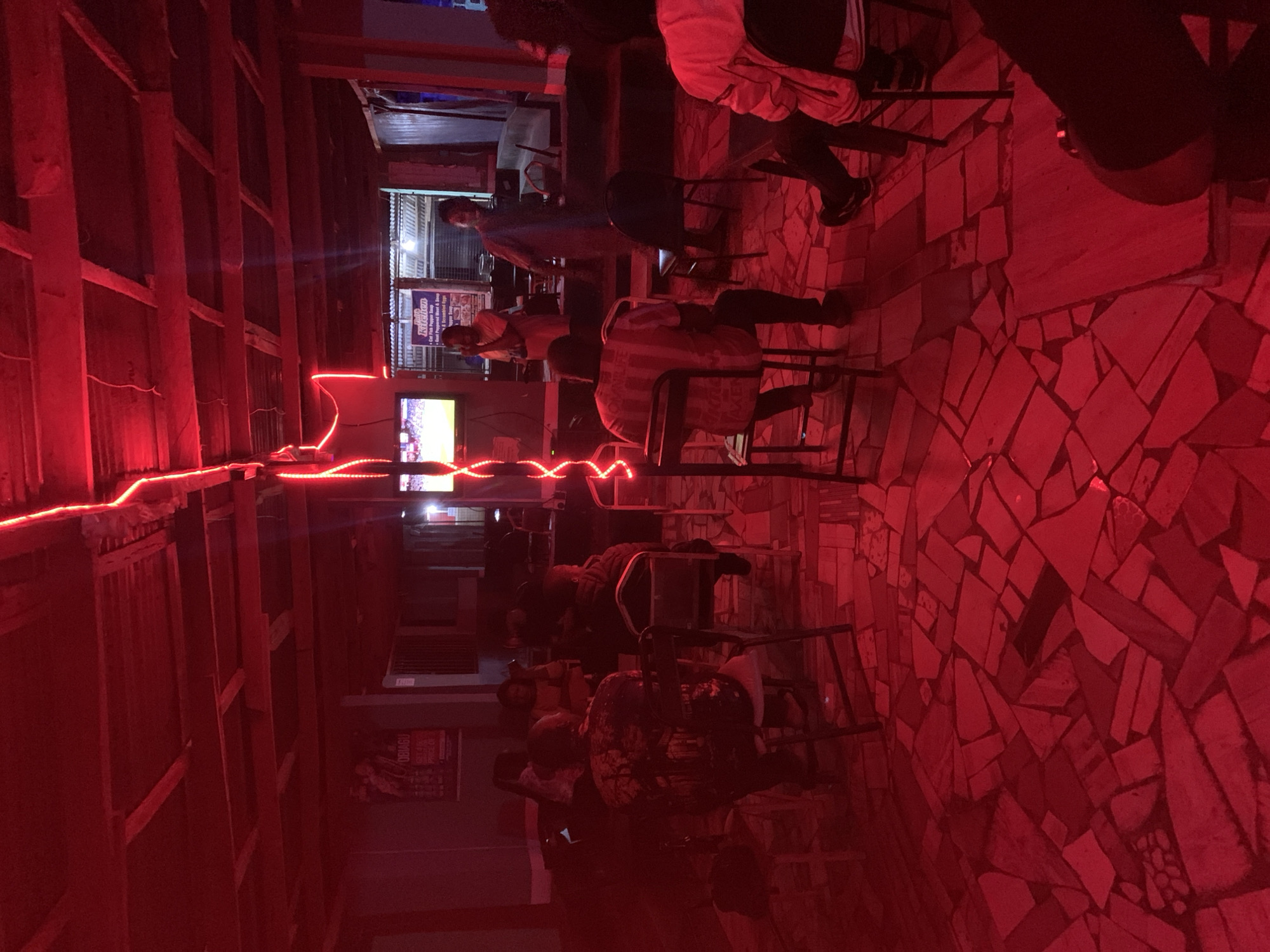 The image shows the interior of a bar in New Stadium, Nigeria. Red lighting pervades the scene. Patrons sit on stools at the bar, some watching a TV screen. The walls appear to be constructed of wood planks. The floor is decorated with a mosaic tile pattern. A red neon light strip runs along the ceiling. A poster is visible on the wall.