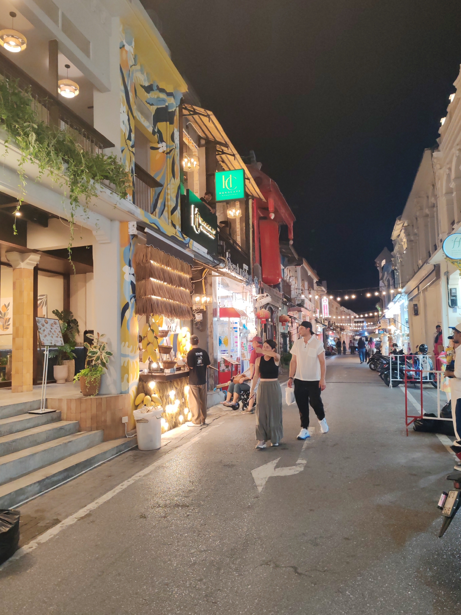 This image captures a vibrant night scene on a narrow, pedestrian-friendly street in Phuket City Municipality, Thailand. The time of day is evening, with a dark sky overhead and the street brightly illuminated by numerous artificial light sources. String lights are strung across the street, and various shops glow with interior and signage lighting.
The street is flanked by multi-story buildings exhibiting different architectural styles. On the left, a white building features a balcony adorned with lush green hanging plants and two spherical pendant lights. Adjacent to it, a building facade is decorated with a distinctive mural in shades of yellow, blue, and white. Below this mural, a shop stands out with a thatched or straw awning, backlit to highlight its features. A bright green sign for "KONGAPP" with a stylized 'K' is visible above this shop. At the base of this shop, a white bucket holds several yellow, mango-like fruits. A person wearing a black shirt with "BONNIE" written on the back is seen standing near this shop. On the right side of the street, buildings showcase classic Sino-Portuguese colonial architecture with white facades, arched windows, and decorative elements.
Numerous people are enjoying the evening atmosphere. In the foreground, a couple walks towards the viewer; the woman wears a dark sleeveless top and light wide-leg pants, while the man is dressed in a white short-sleeved shirt and black pants, carrying a white bag. Other pedestrians can be seen further down the street, some strolling and others gathered near shops. Motorcycles are parked along the right side of the street. A white arrow painted on the asphalt road indicates a forward direction. The overall scene conveys a lively and inviting ambiance, characteristic of a night market or a popular evening promenade.