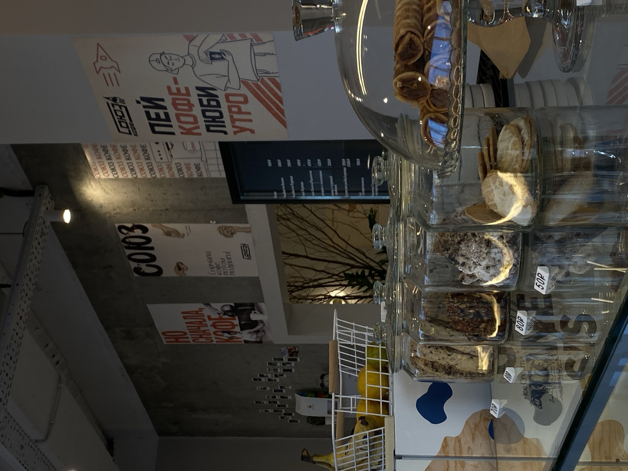 The image depicts the interior of a cafe, likely in the city of Agronom, Russia, as suggested by the posters on the wall. The cafe has a modern, industrial aesthetic with concrete walls and exposed ceiling elements. On the left, a white wire basket contains bananas, lemons, and what appear to be limes. Behind the fruit, there are small framed photographs adhered to the wall.
Dominating the background are several large posters. One poster, featuring bold red and black text, reads "HO CHAYAMA, KOFRE," with a depiction of a dog and a mug. Another poster, with a red rocket symbol, has the text "SOYUZ" and "PEI KOFRE - LYUBI UTRO," suggesting a coffee-related theme. A digital menu board is visible on a stand to the right of the posters, displaying various items and prices in rubles.
In the foreground, a glass display counter showcases baked goods. Several large glass jars are filled with different kinds of cookies and dry snacks. To the right, a tiered glass stand holds a stack of rolled wafer cookies, possibly filled with chocolate. The reflection on the glass counter reveals a cow graphic and the words "ice cream." Price tags with "70P," "80P," and "50P" are visible in front of the jars, indicating pricing in rubles. The lighting in the cafe is somewhat dim, with spotlights directed at specific areas, creating highlights and shadows. The overall impression is a trendy and inviting coffee shop.