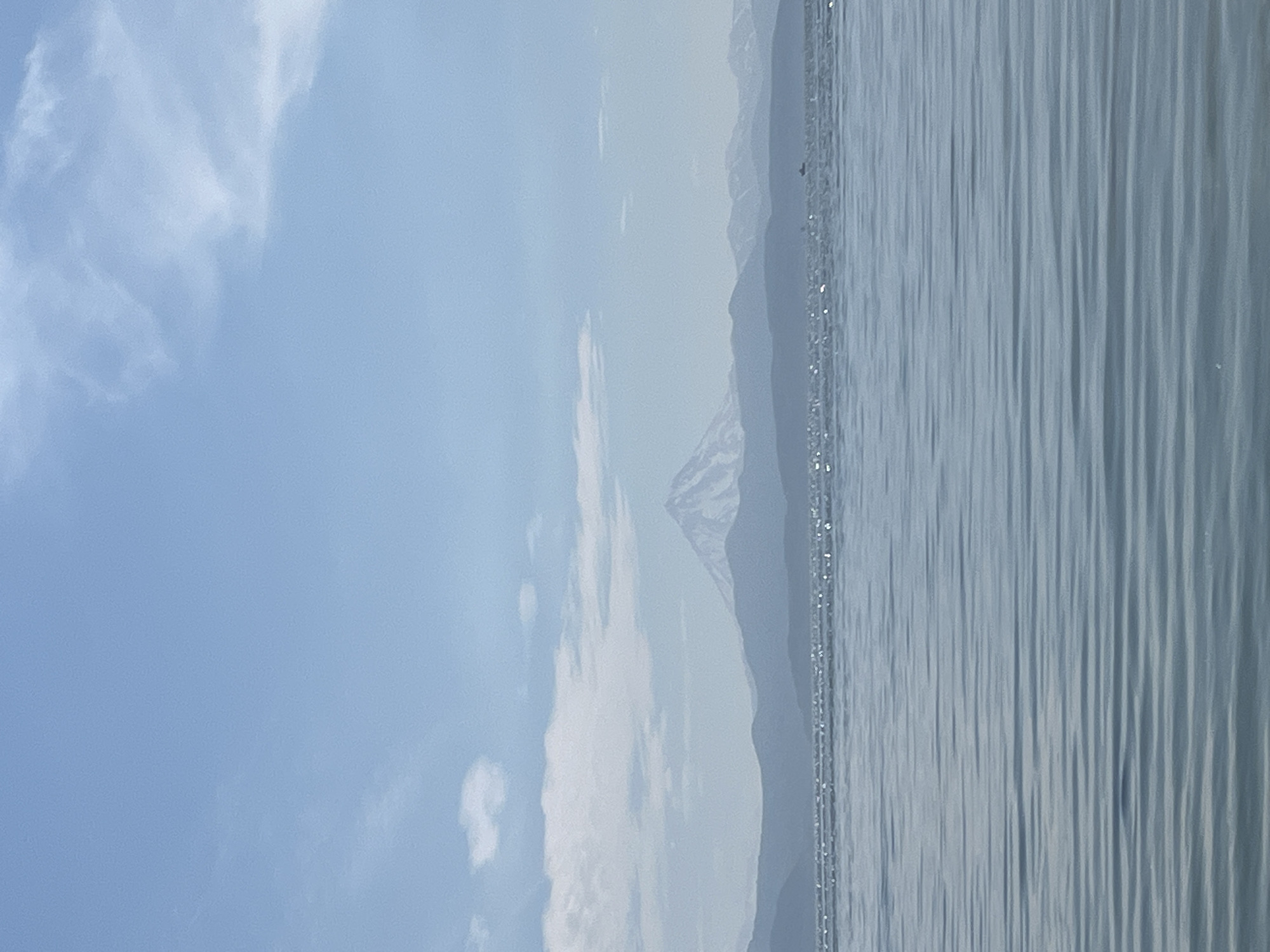 The image shows a wide, tranquil body of water in the foreground, with gentle ripples reflecting the bright sky. In the distance, a range of hazy mountains forms a backdrop, the tallest of which is a snow-capped volcanic peak. The sky above is a clear, pale blue, with scattered white clouds. The overall impression is one of a serene, natural landscape. This scene is likely taken from the coast or a boat in Avacha Bay, looking towards the volcanoes of the Kamchatka Peninsula near Petropavlovsk-Kamchatsky, Russia.